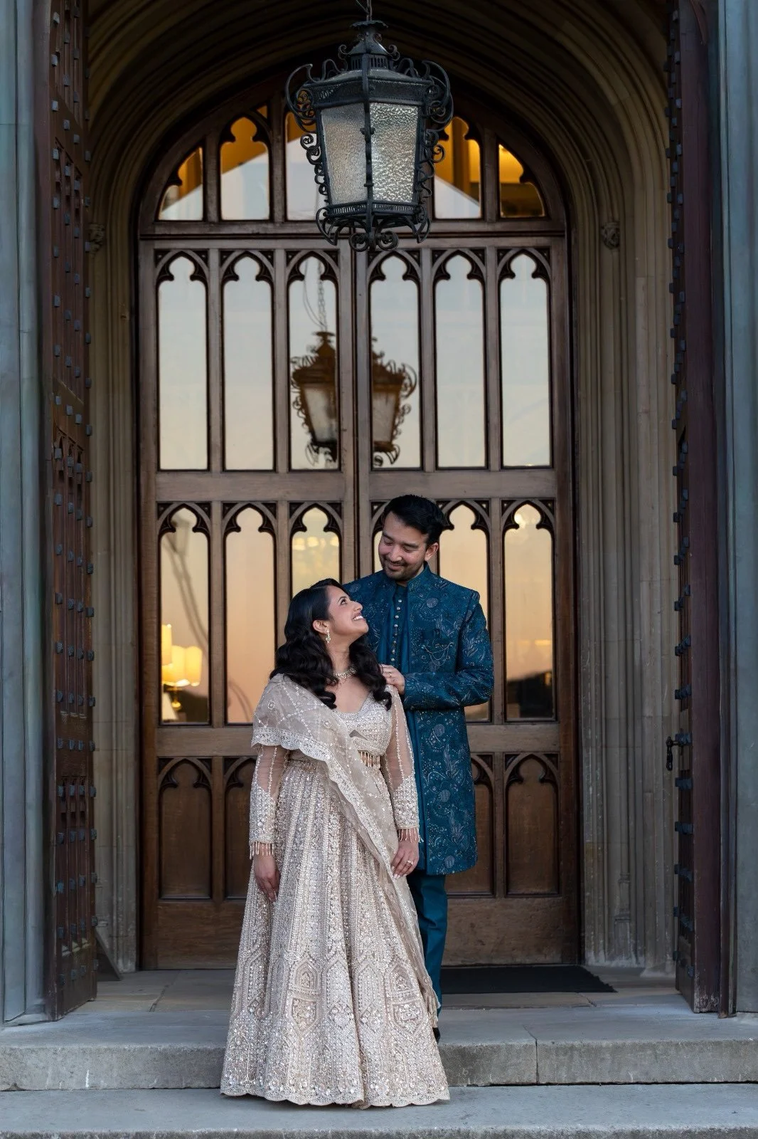 A South Asian couple in traditional attire standing on a stone porch in front of an arched wooden door with glass panels, smiling at each other during sunset.