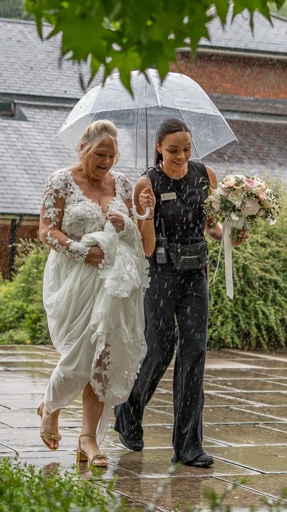 A bride and a woman holding an umbrella walking in the rain, the bride smiling and wearing a white wedding dress, while the other woman, dressed in black, holds a bouquet of flowers.