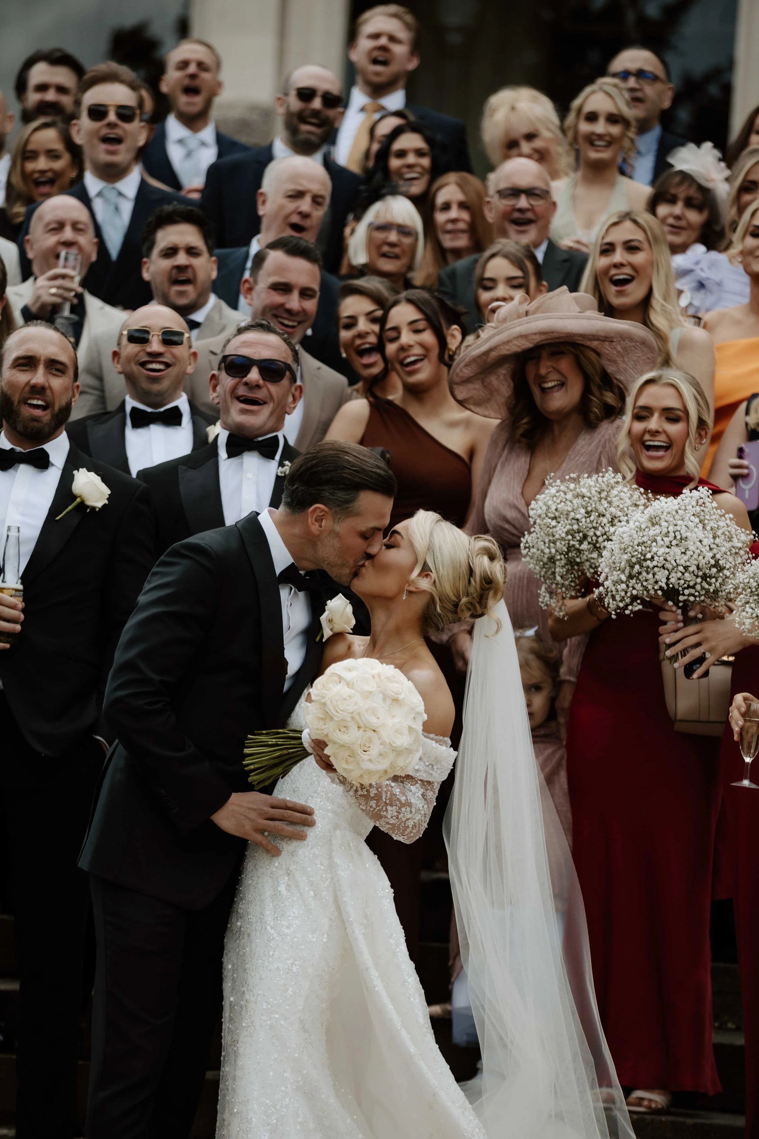 A wedding couple kisses, surrounded by a joyful crowd. The bride is in a white gown holding a bouquet of white roses, and the groom is in a black tuxedo. The crowd includes men and women in formal attire, some wearing sunglasses, smiling, and celebra