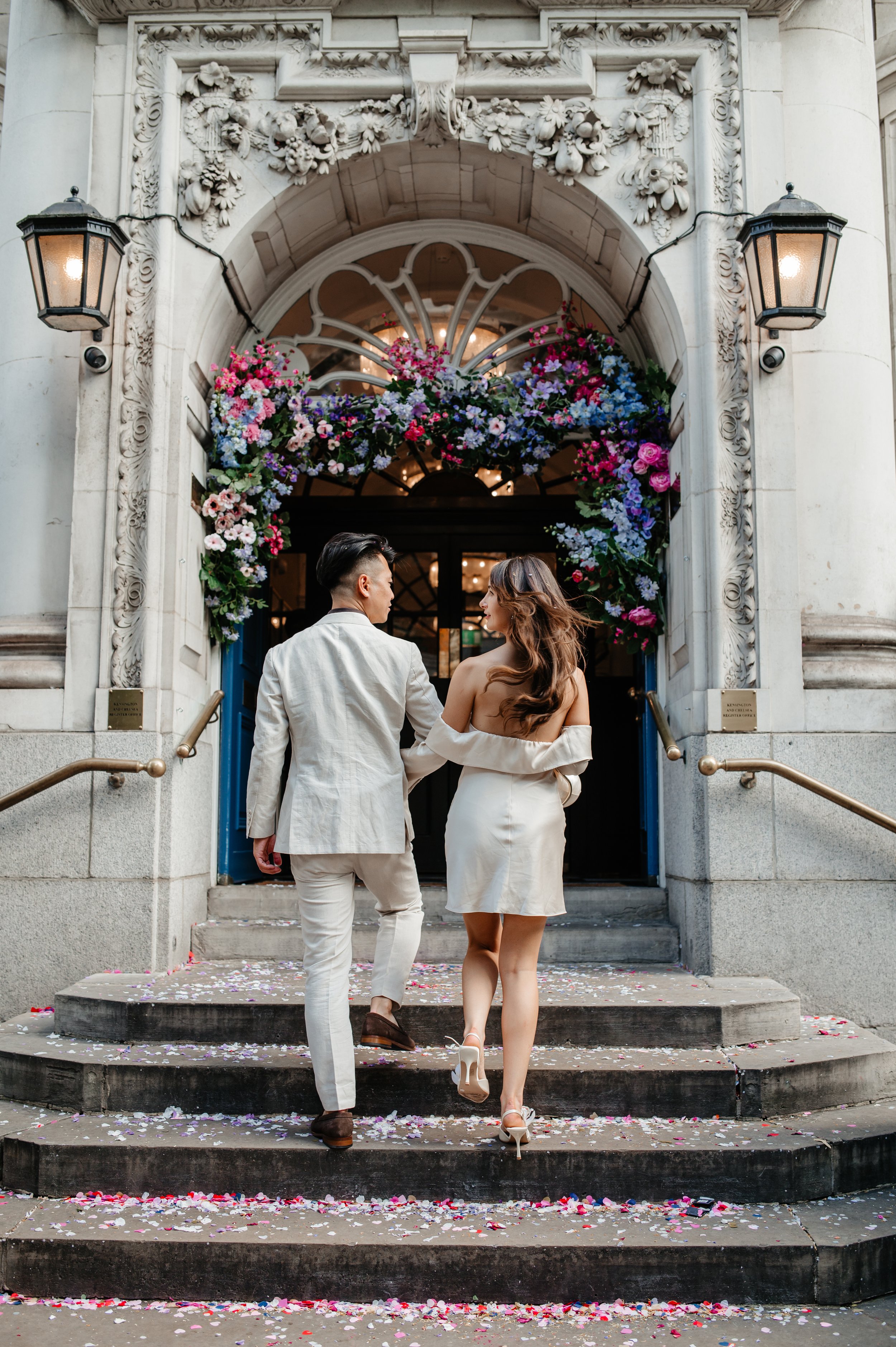 Couple in wedding attire walking up steps decorated with flower petals, ascending into a building with ornate stonework and floral decorations at the entrance.