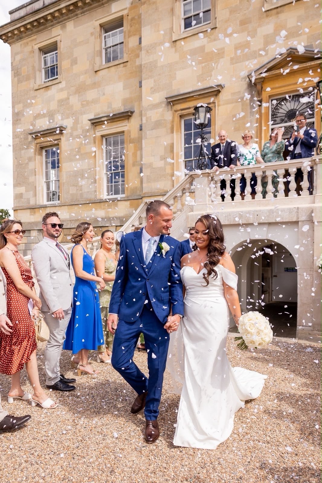 A wedding celebration with a bride and groom walking hand-in-hand, surrounded by guests outside a historic stone building, with confetti falling around them.