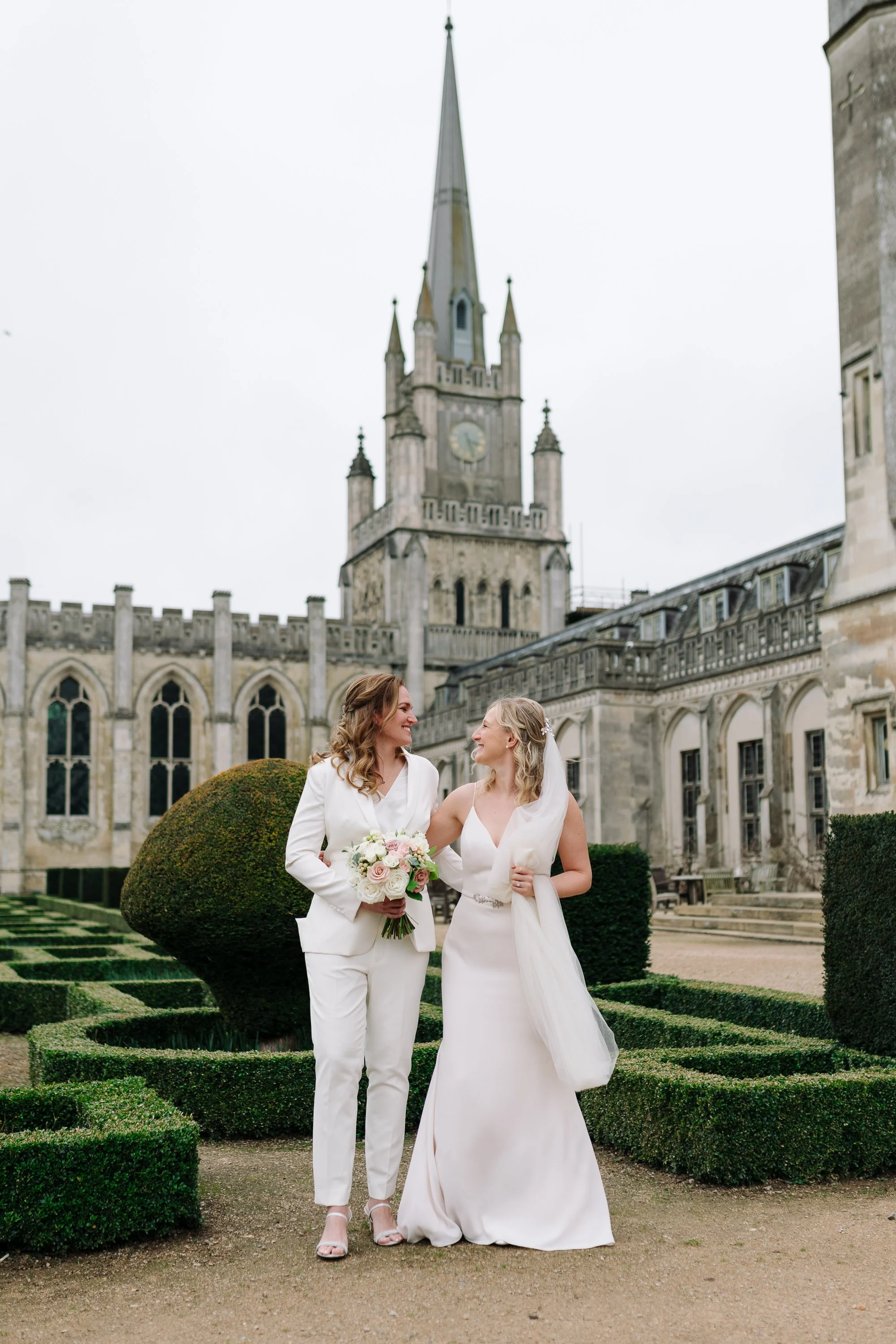 Two women in wedding attire standing close together in a formal garden with trimmed hedges, with a historic stone building and a clock tower in the background. They are smiling and looking into each other's eyes, with one woman holding a bouquet of f