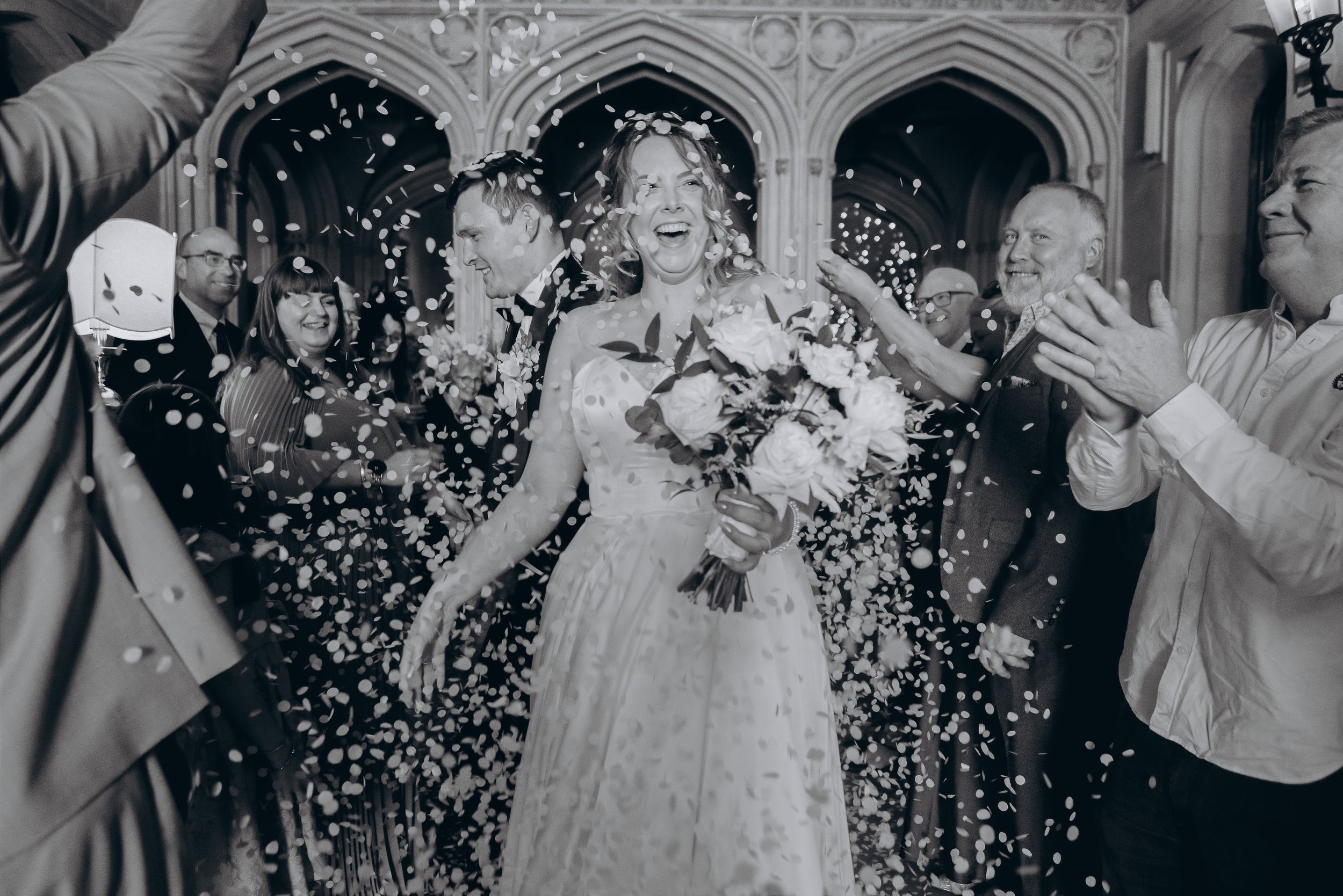 A bride holding a bouquet celebrating with guests in a Gothic-style church as confetti falls around her.