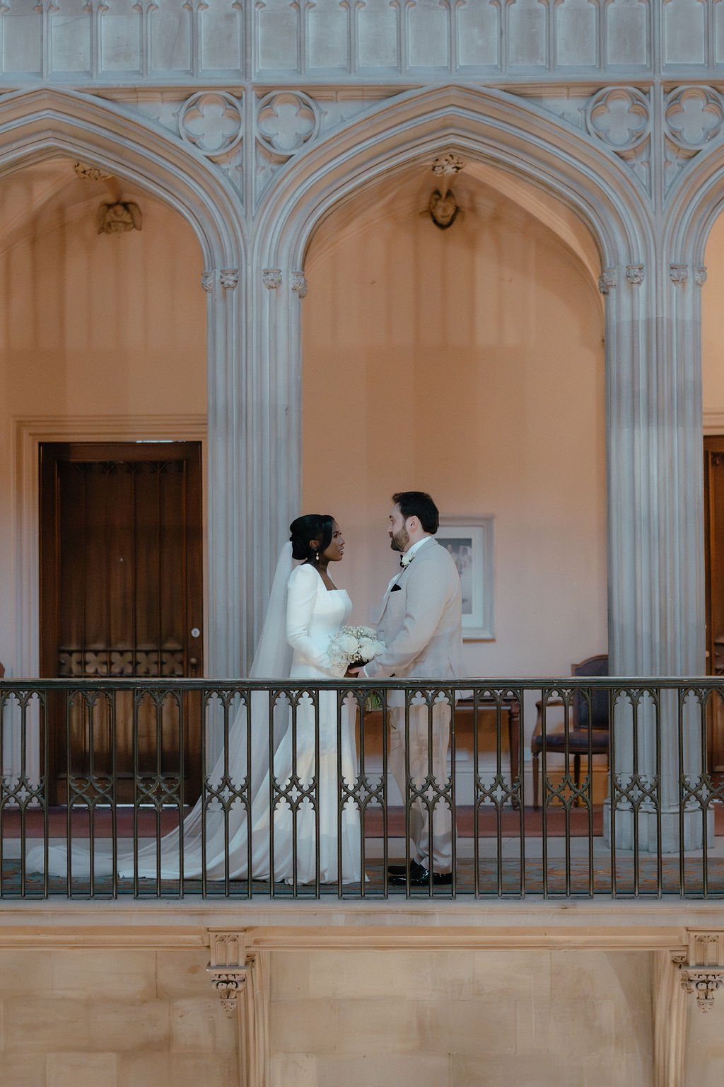 A bride and groom stand facing each other on a balcony, holding hands and gazing into each other's eyes in a historic, ornate building with gothic architectural elements.