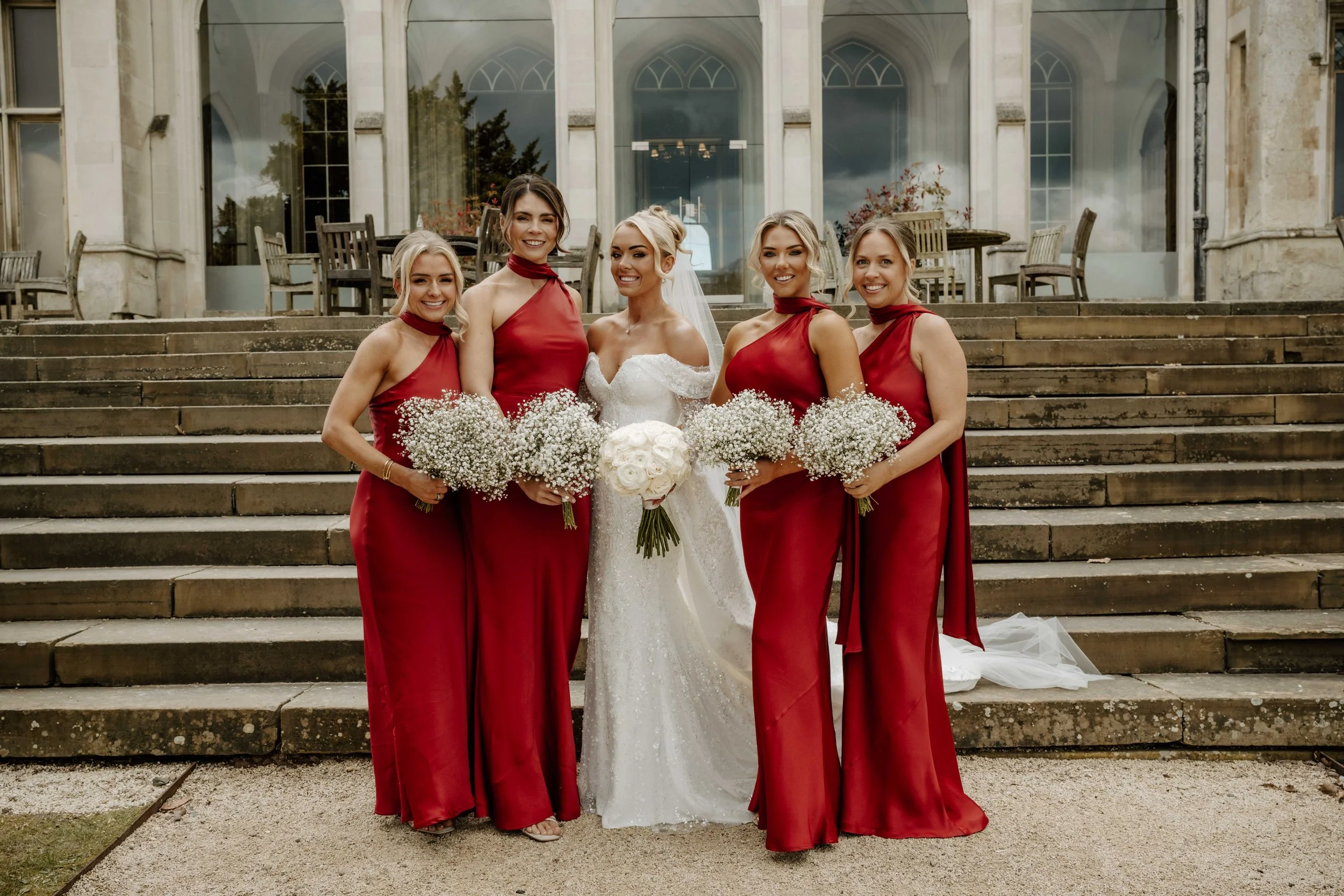 A bride with four bridesmaids standing on stairs in front of a large building with glass doors and windows. The bridesmaids are wearing matching red dresses and holding bouquets of baby's breath flowers. The bride is in a white wedding gown with a ve