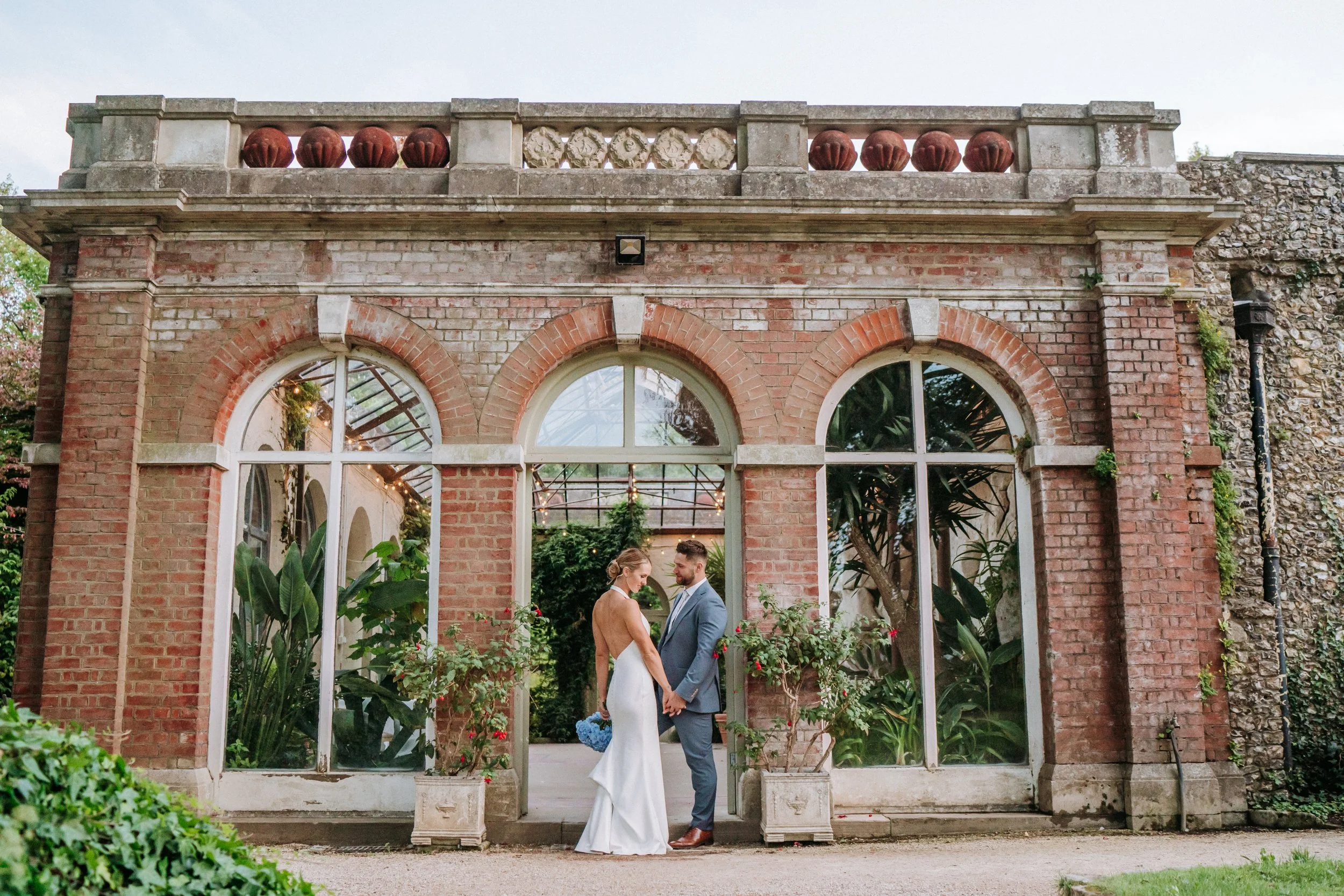 A bride and groom holding hands and facing each other, inside a glass-enclosed greenhouse with brick walls, green plants, and string lights.