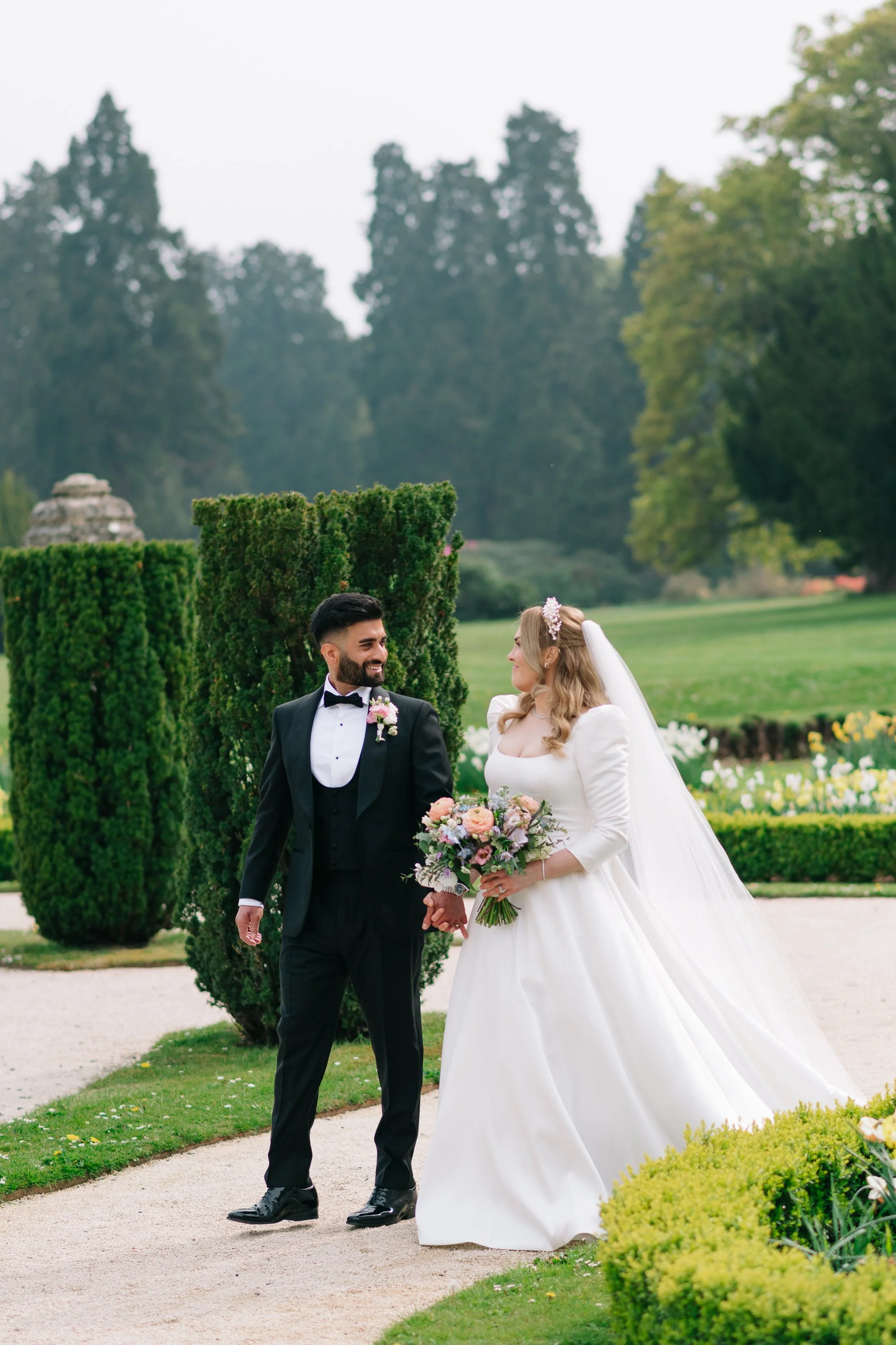 A bride and groom are standing outdoors in a garden setting, smiling at each other during their wedding. The groom is wearing a black tuxedo with a bow tie, and the bride is in a white wedding gown holding a bouquet of flowers.