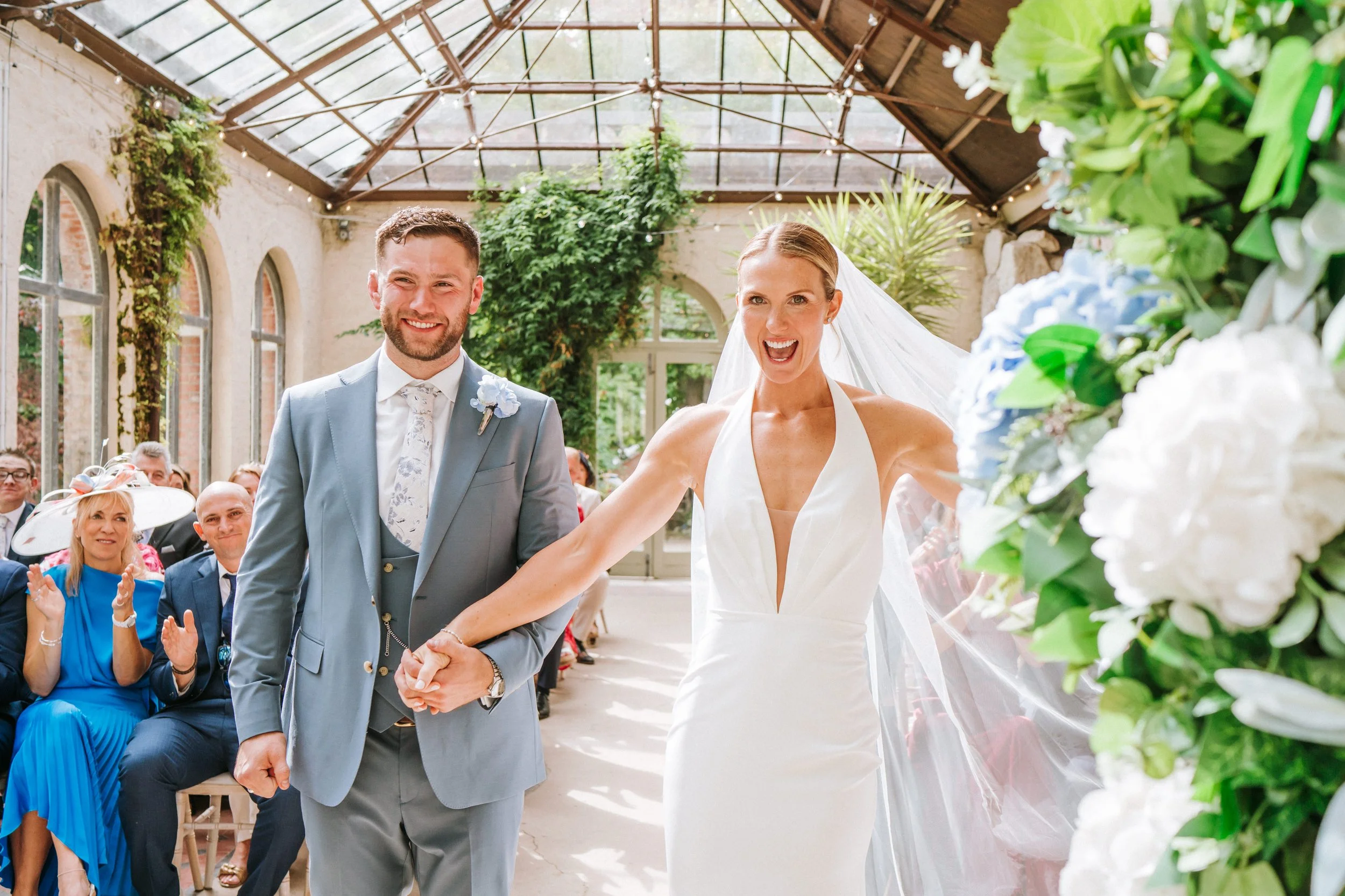 A bride and groom holding hands during their wedding ceremony in a conservatory or greenhouse with glass ceiling, surrounded by guests.