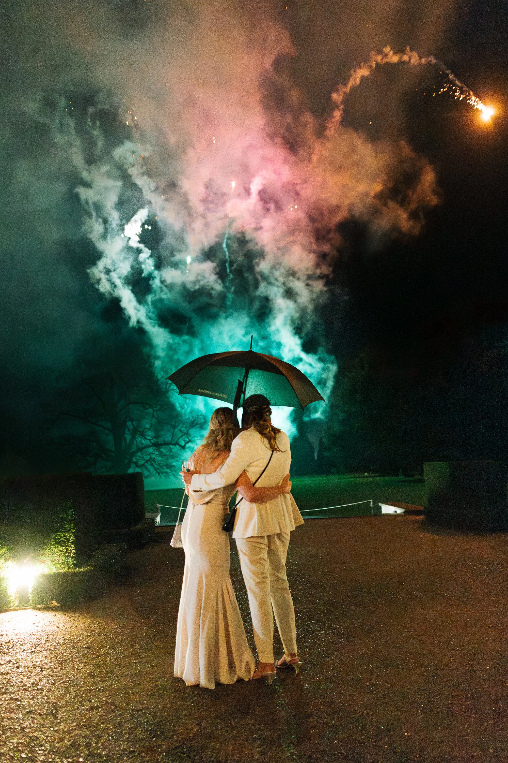 Two women in elegant white dresses stand close together at night, sharing an umbrella, with fireworks illuminating the sky in the background.