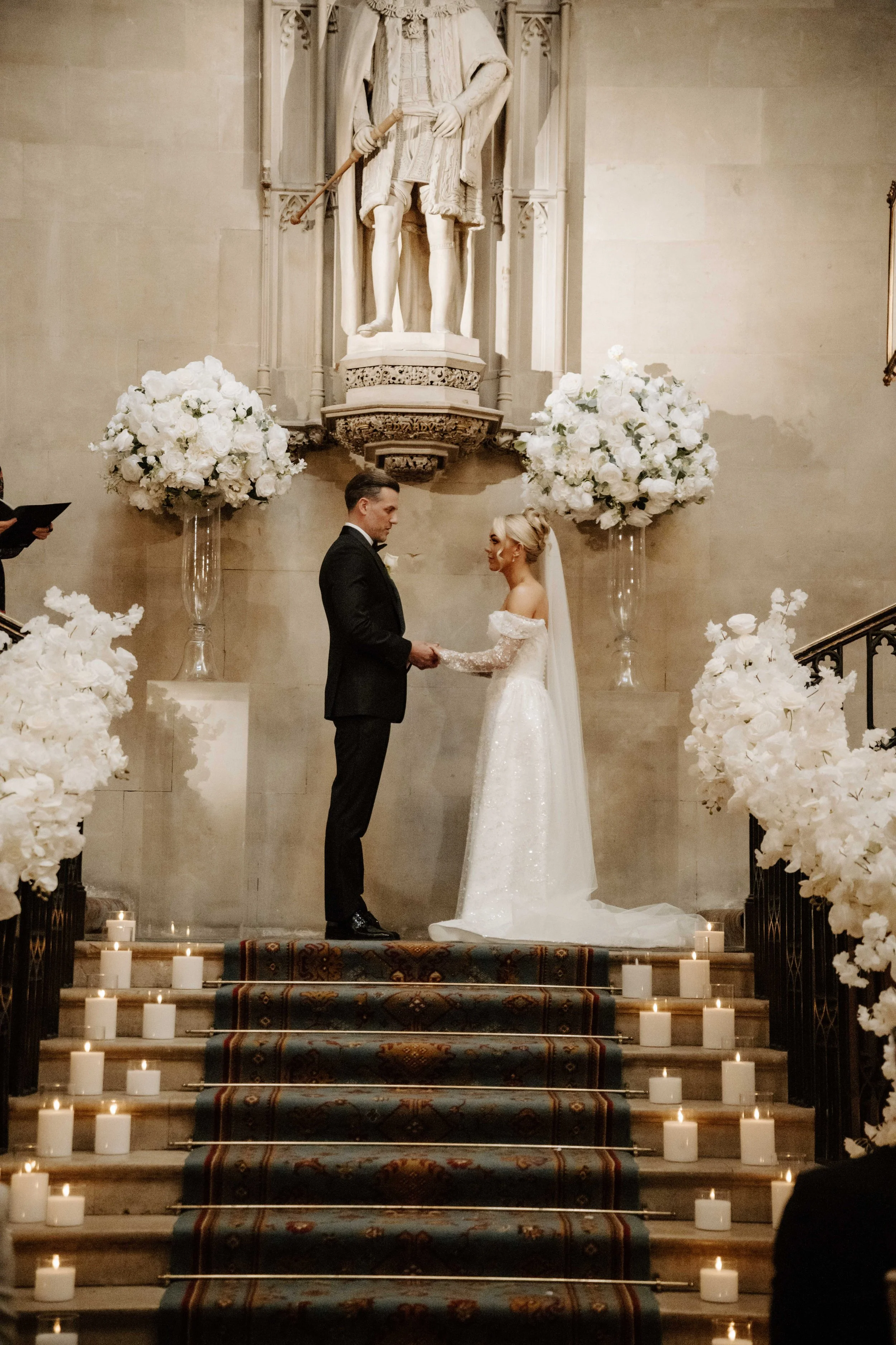 A bride and groom exchanging vows at the top of a decorated staircase in a church, surrounded by large white floral arrangements and candles.