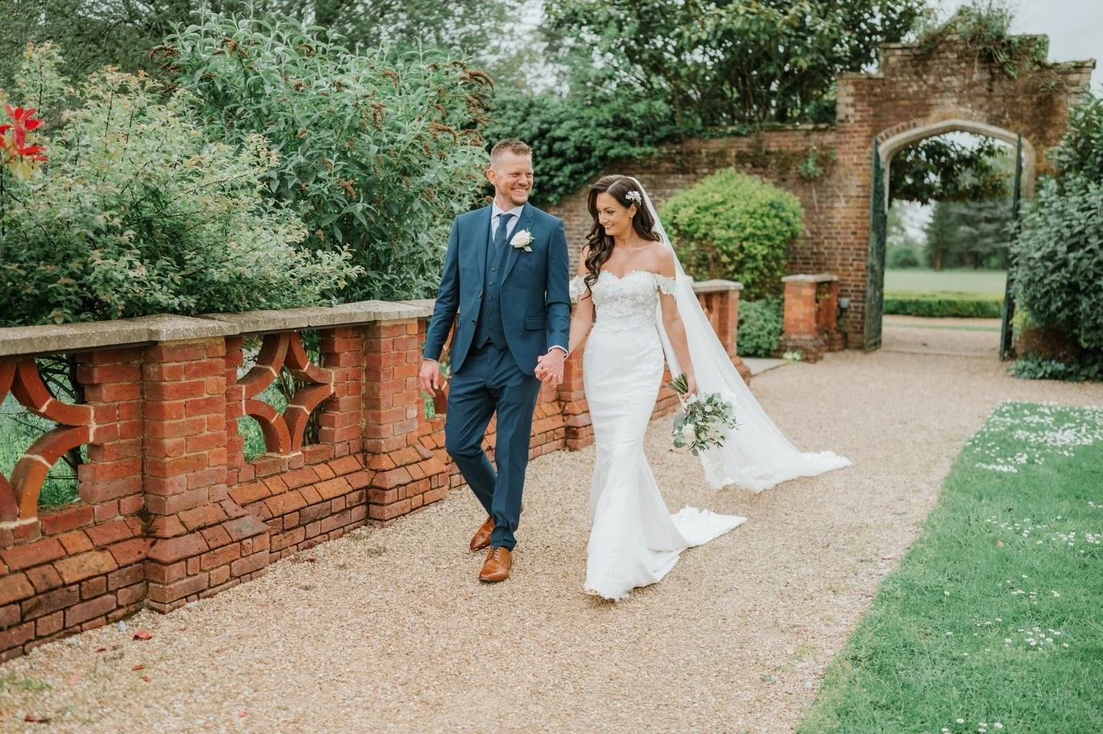 A bride and groom walking hand in hand outdoors near a brick archway, surrounded by green bushes and trees on a gravel path, during their wedding day.