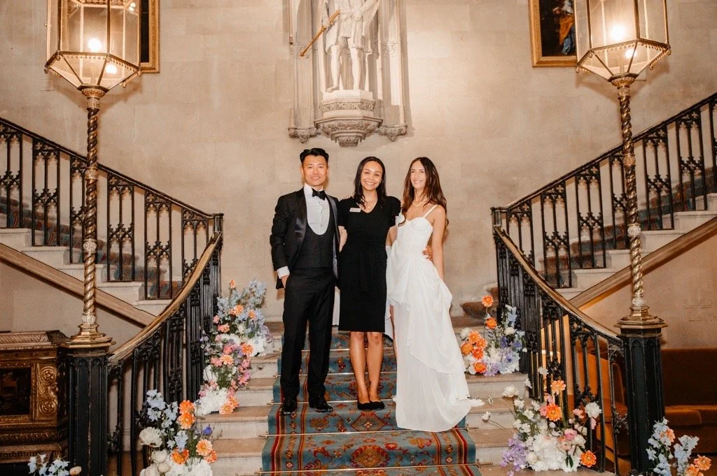 A wedding couple and a woman stand on a staircase with floral arrangements, inside a historic building with ornate railings and large lamps.