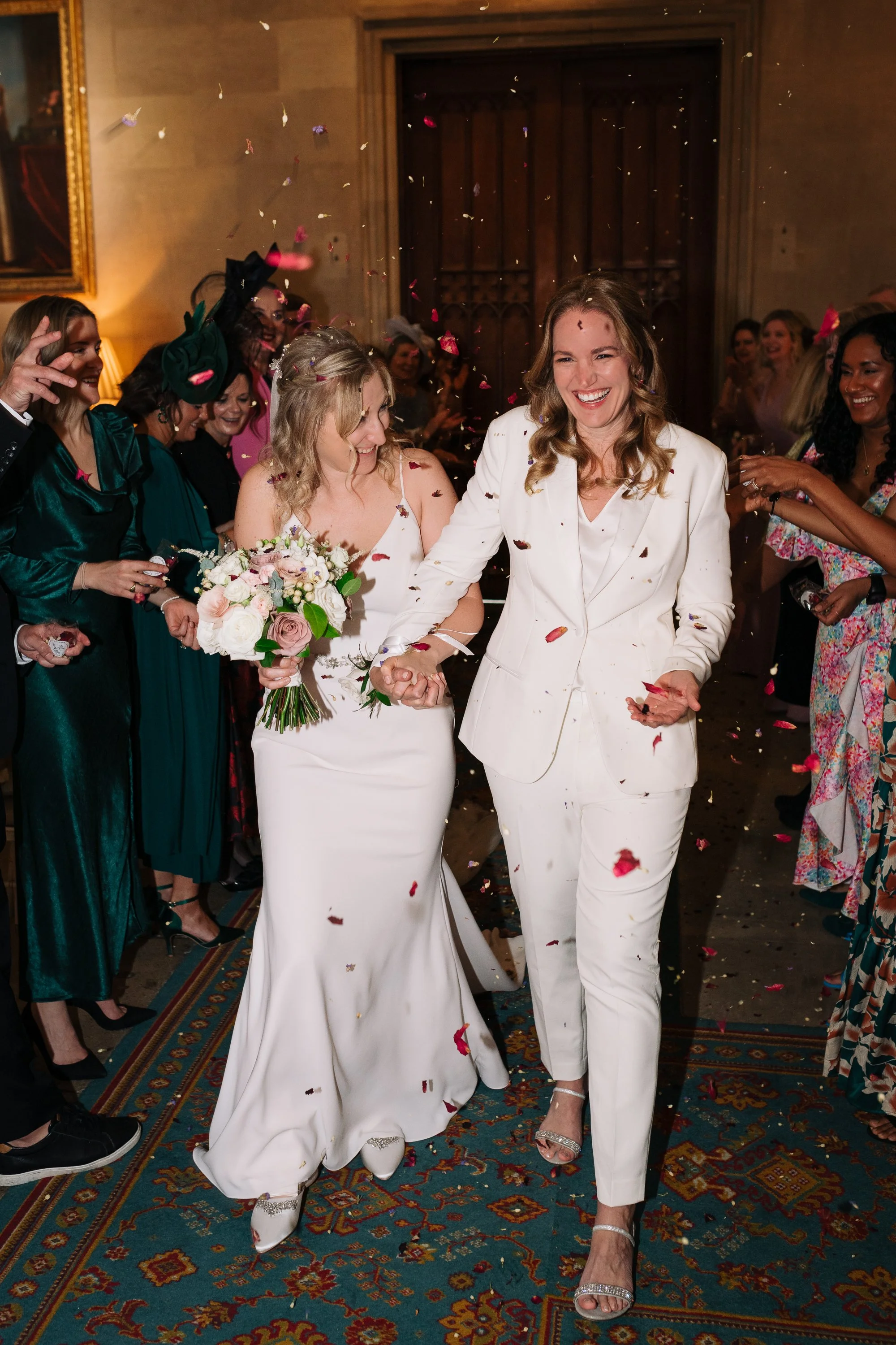 Two women in white dresses, one with a bouquet, holding hands and smiling as they walk through a party with guests throwing confetti.