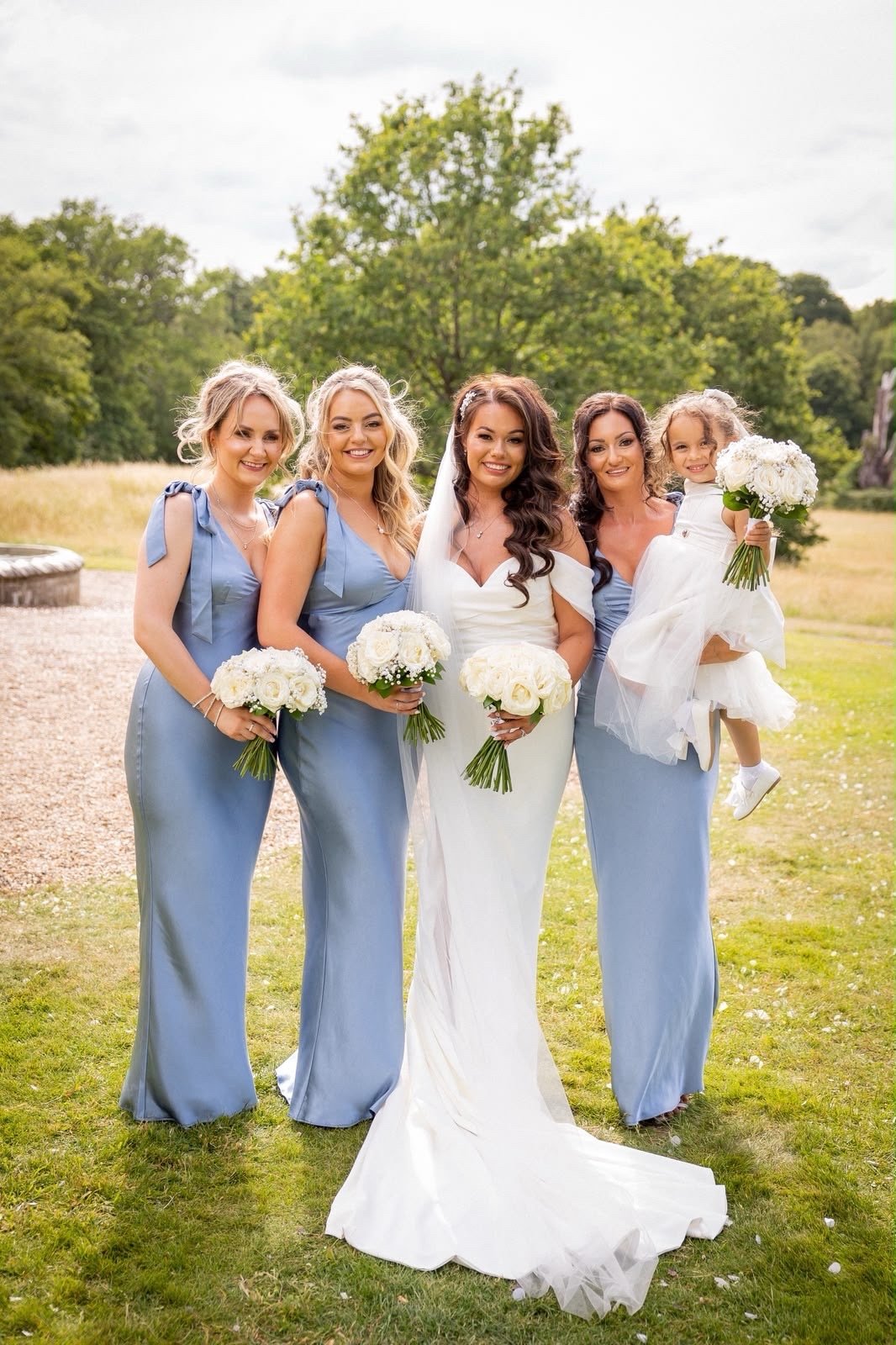 A bride and four bridesmaids standing outdoors on grass, holding bouquets of white flowers. The bride is in a white wedding gown, the bridesmaids are in light blue dresses. One of the bridesmaids is holding a young girl in a white dress, also holding