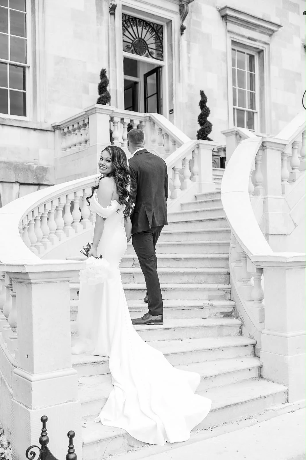A bride in a white wedding dress holding a bouquet stands on the staircase of a large, elegant building, smiling at the camera. A groom in a dark suit is behind her, facing away. The building has large windows, decorative columns, and a clock above t