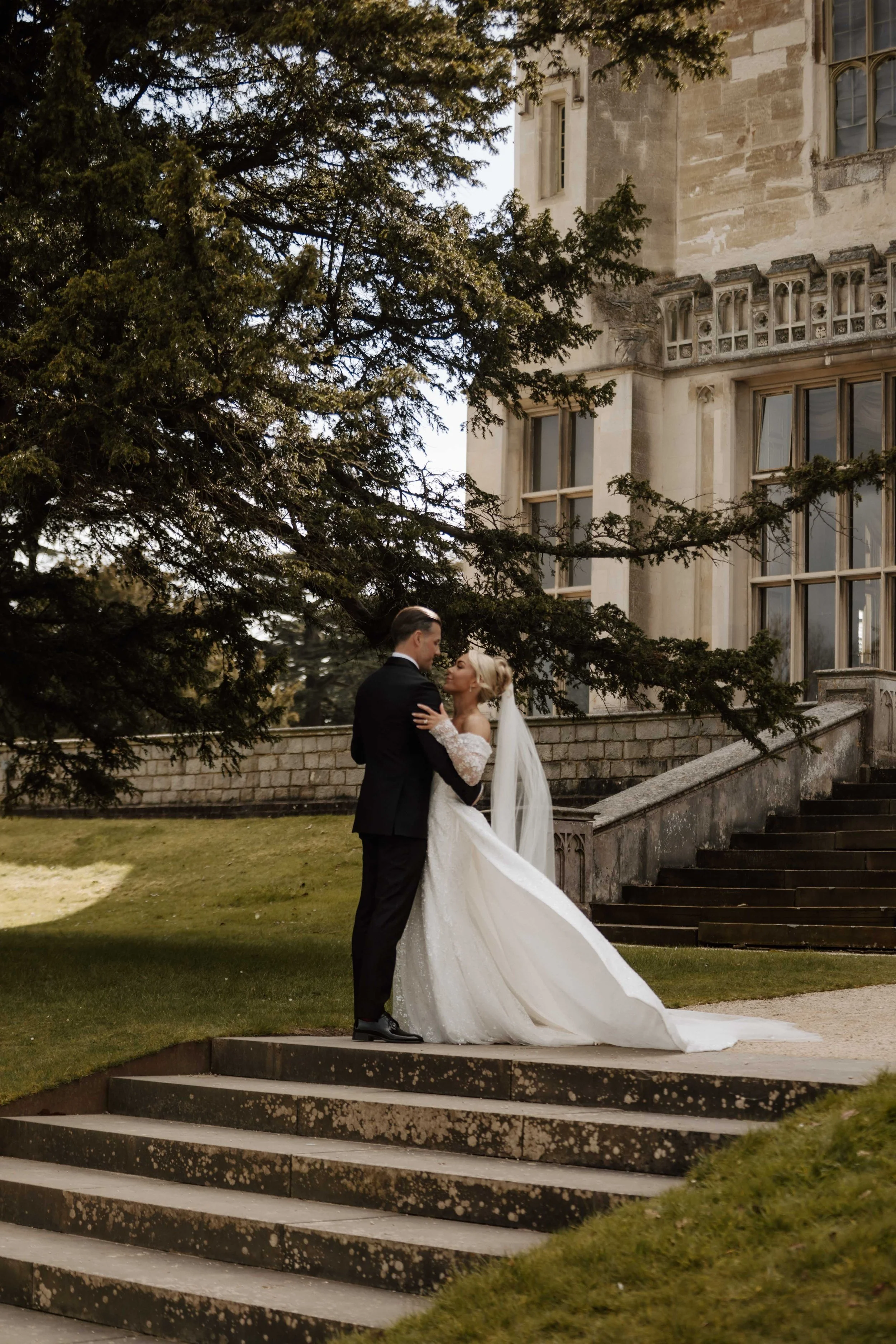 A bride and groom standing on stone steps outside a historic stone building, embracing under a large pine tree.