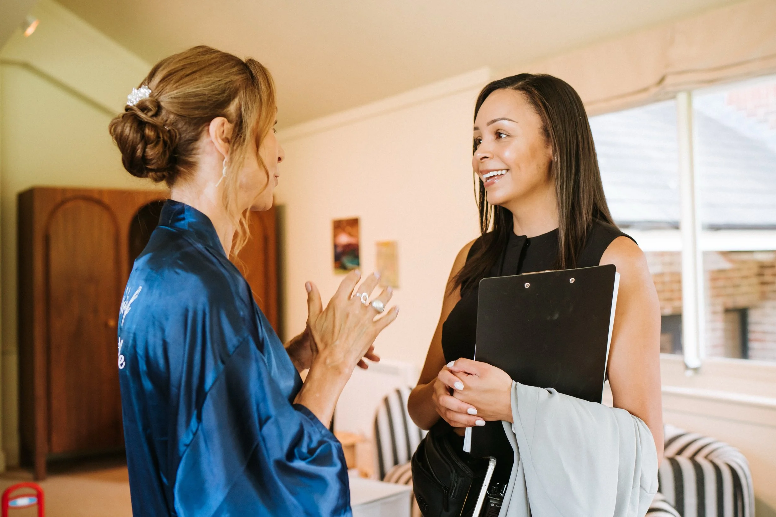 Two women talking indoors, one holding a clipboard and the other wearing a blue jacket with pearl hair accessories.