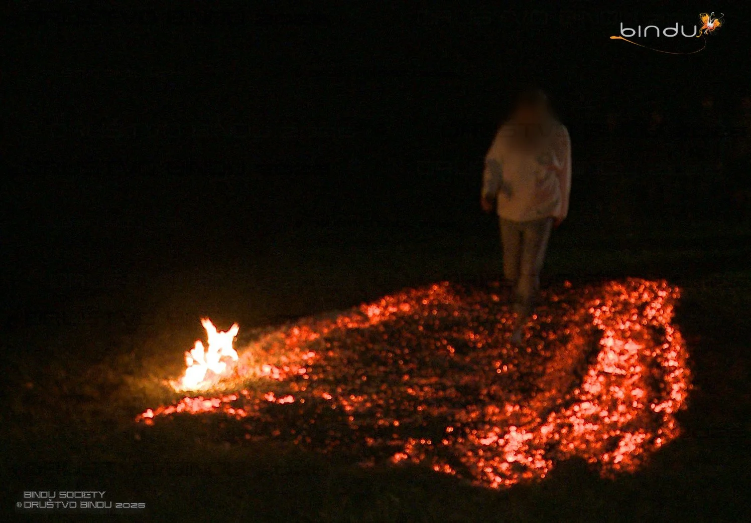 Person walking on a path of burning embers at night, with a small flame at the start of the trail.