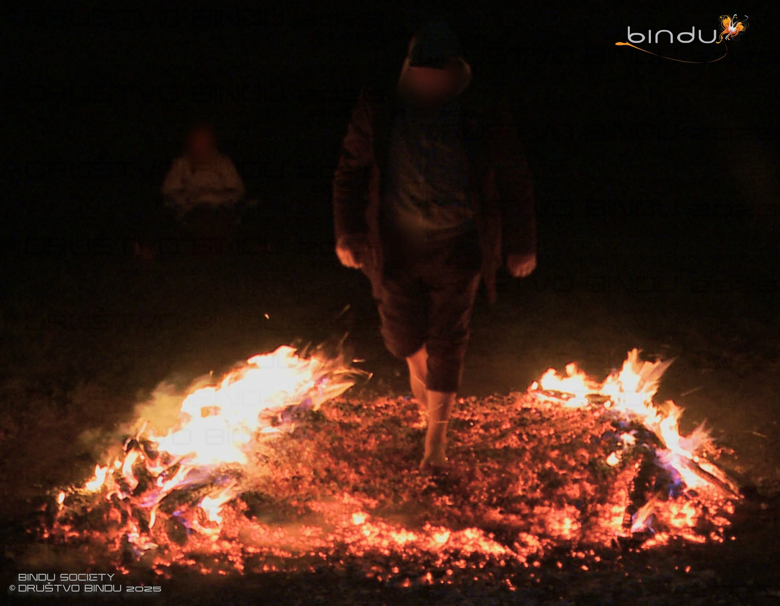 A person standing on a fire-lit ground at night, with their face obscured by a hood, surrounded by small fires and glowing embers.