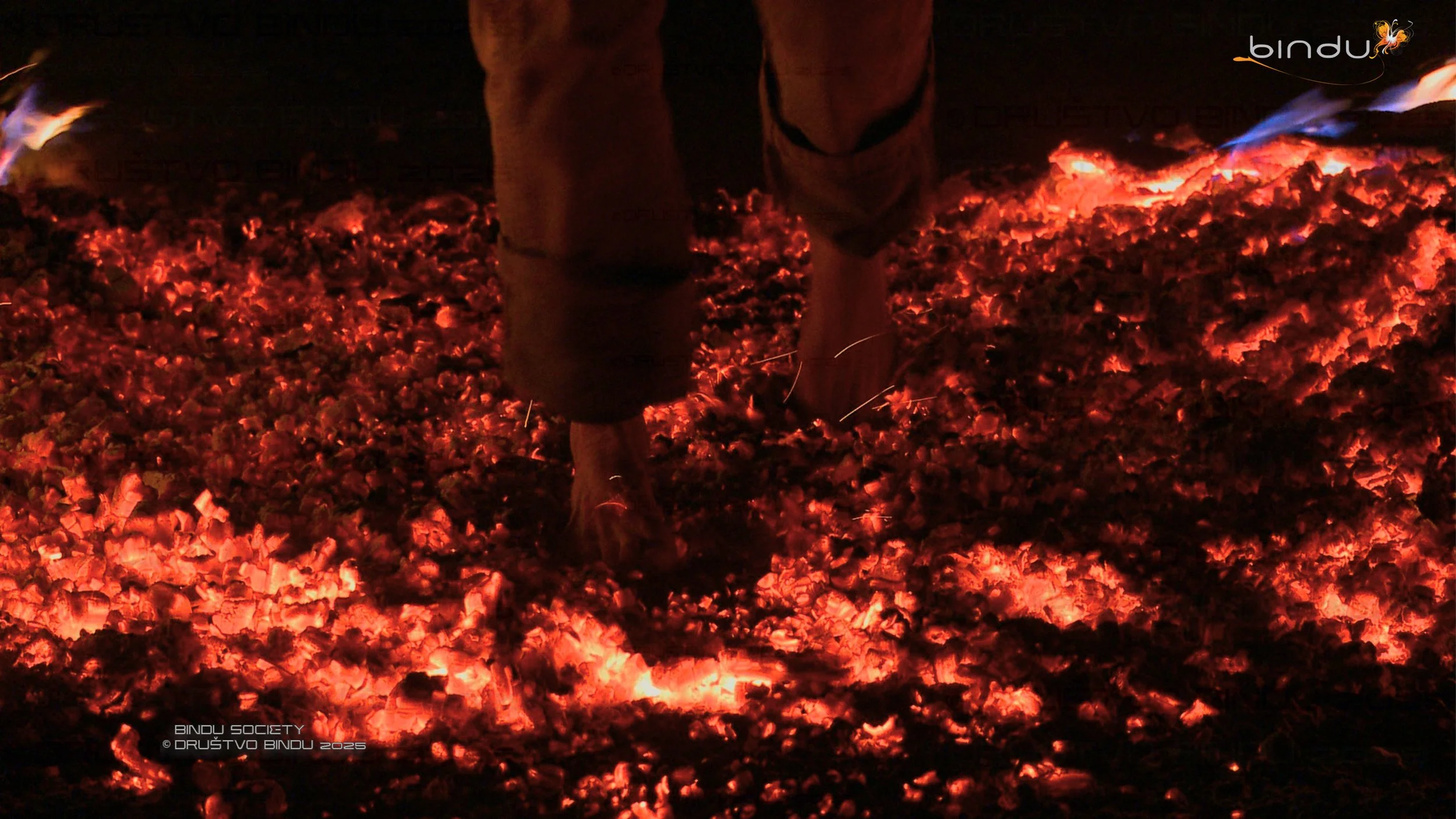 Close-up of a person in brown boots and beige pants standing on glowing red and orange volcanic embers in darkness, with blue flames in the background.