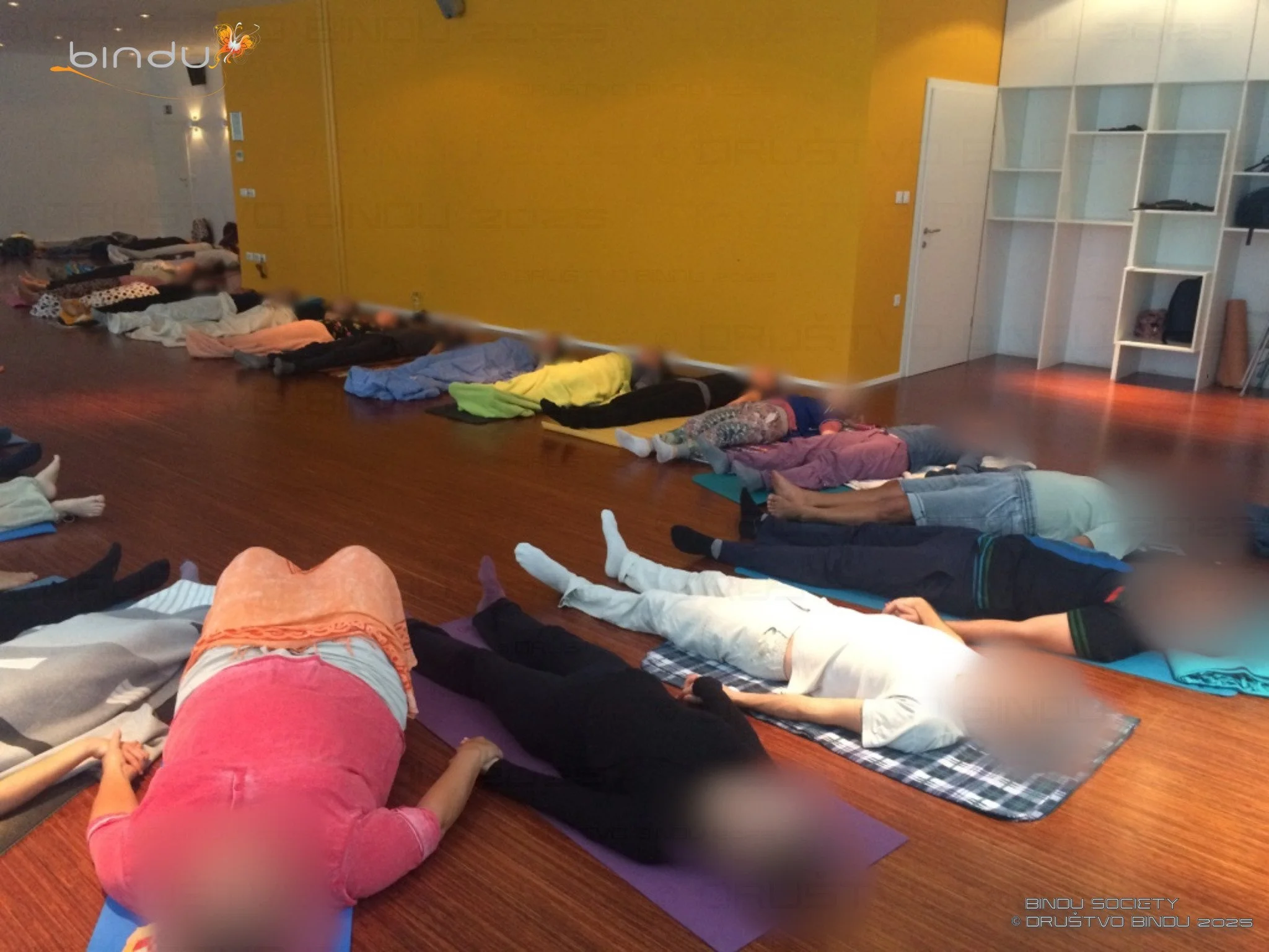 Group of people lying on yoga mats in a room, practicing a relaxation or meditation pose with eyes closed.