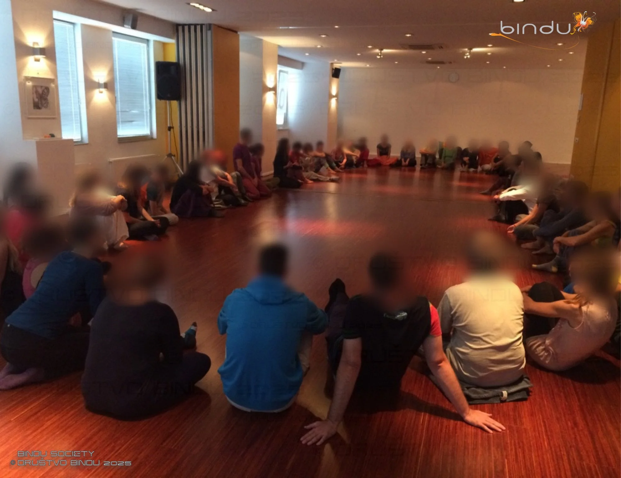 A group of people are sitting and kneeling on the floor of a room, engaged in a yoga or meditation session. The room has large windows, soft lighting, and a wooden floor, with a person in the front performing a yoga pose.