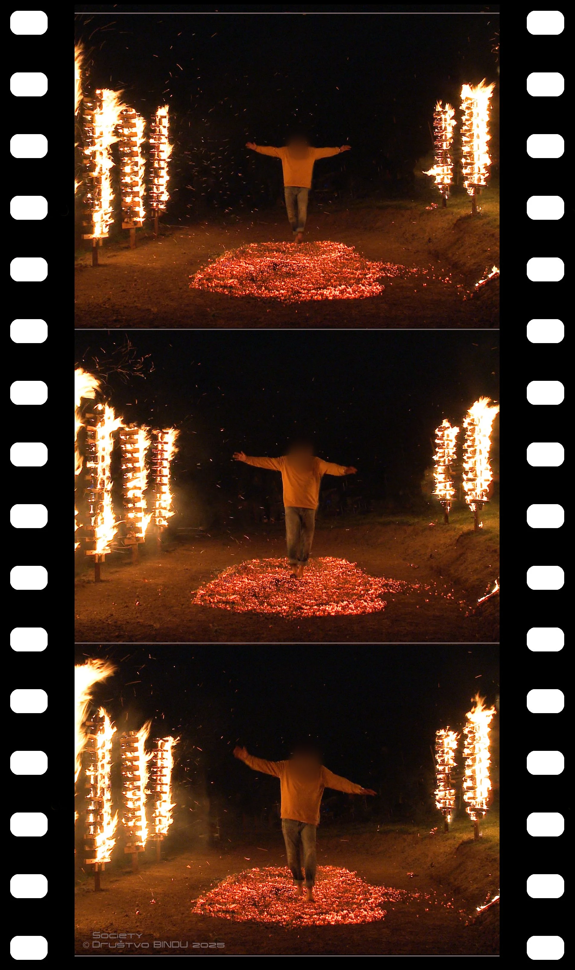 A person stands with arms outstretched in the center of a path illuminated by ground fires, with vertical flames on either side of the path against a dark night background, creating a dramatic scene.