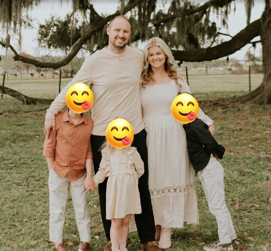 A family of five posing outdoors in a park with large trees in the background. The family members have emojis covering their faces, with two children, a man, a woman, and a young boy.
