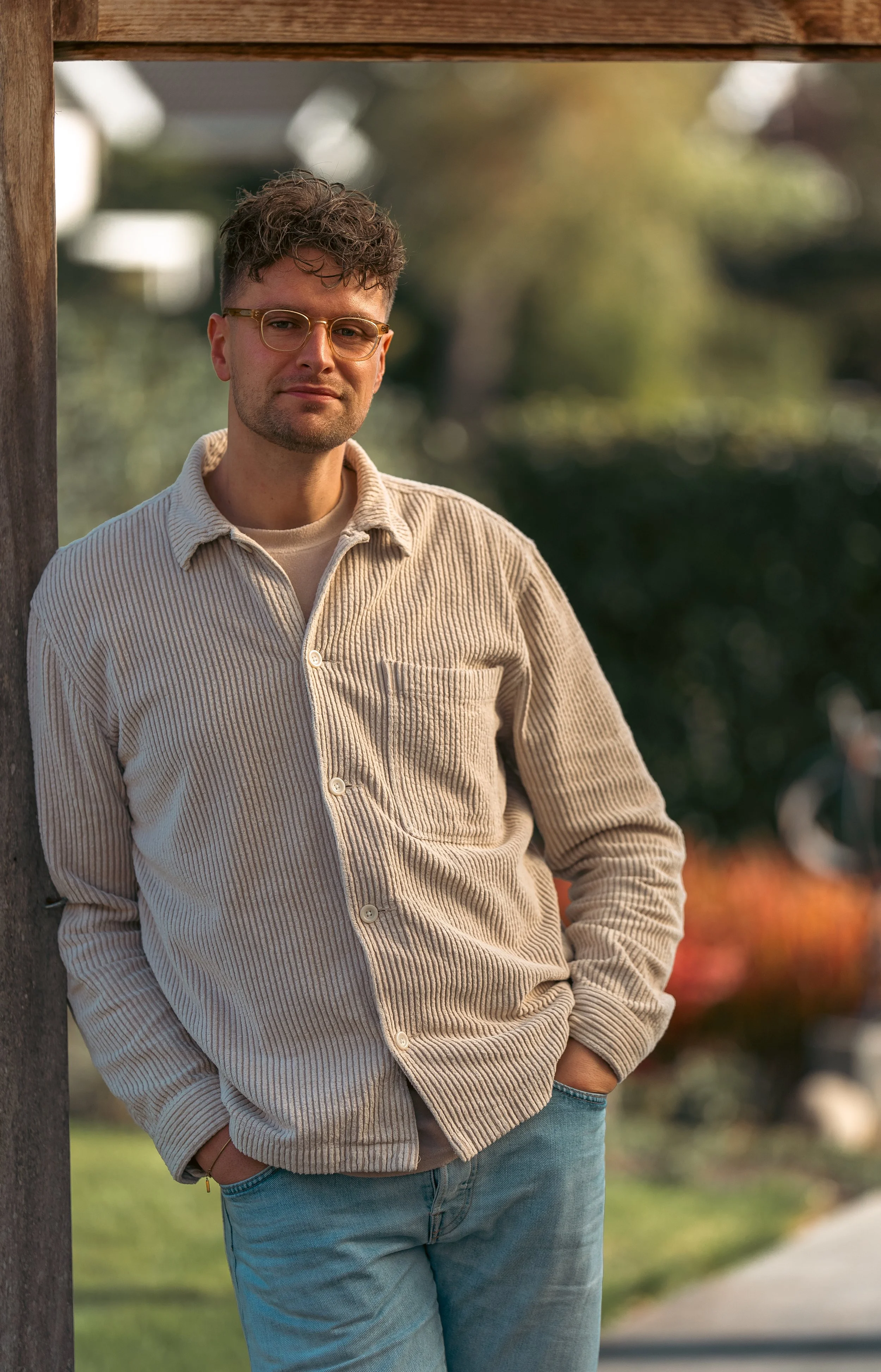 Man leunt tegen houten paal in een park op een zonnige dag, draagt een beige gestreepte jas en blauwe jeans, met een ontspannen uitdrukking.