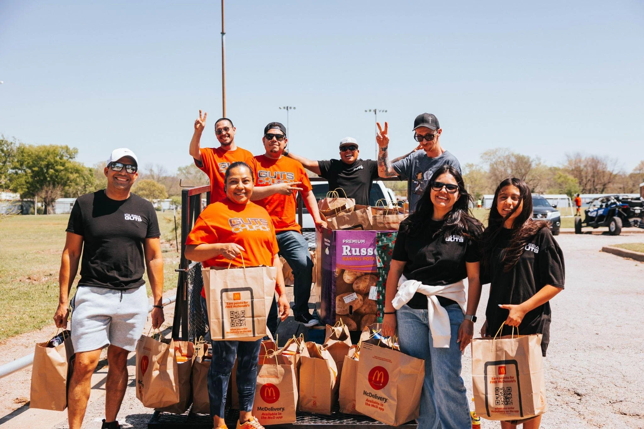 Group of smiling people standing outdoors with shopping bags and boxes for a community event or volunteer activity, some wearing black and orange shirts, with a truck and trees in the background.