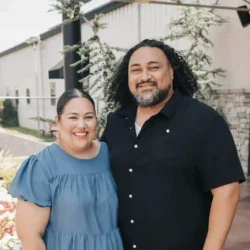A smiling woman and man standing outdoors in front of a building, with trees and a fence in the background.