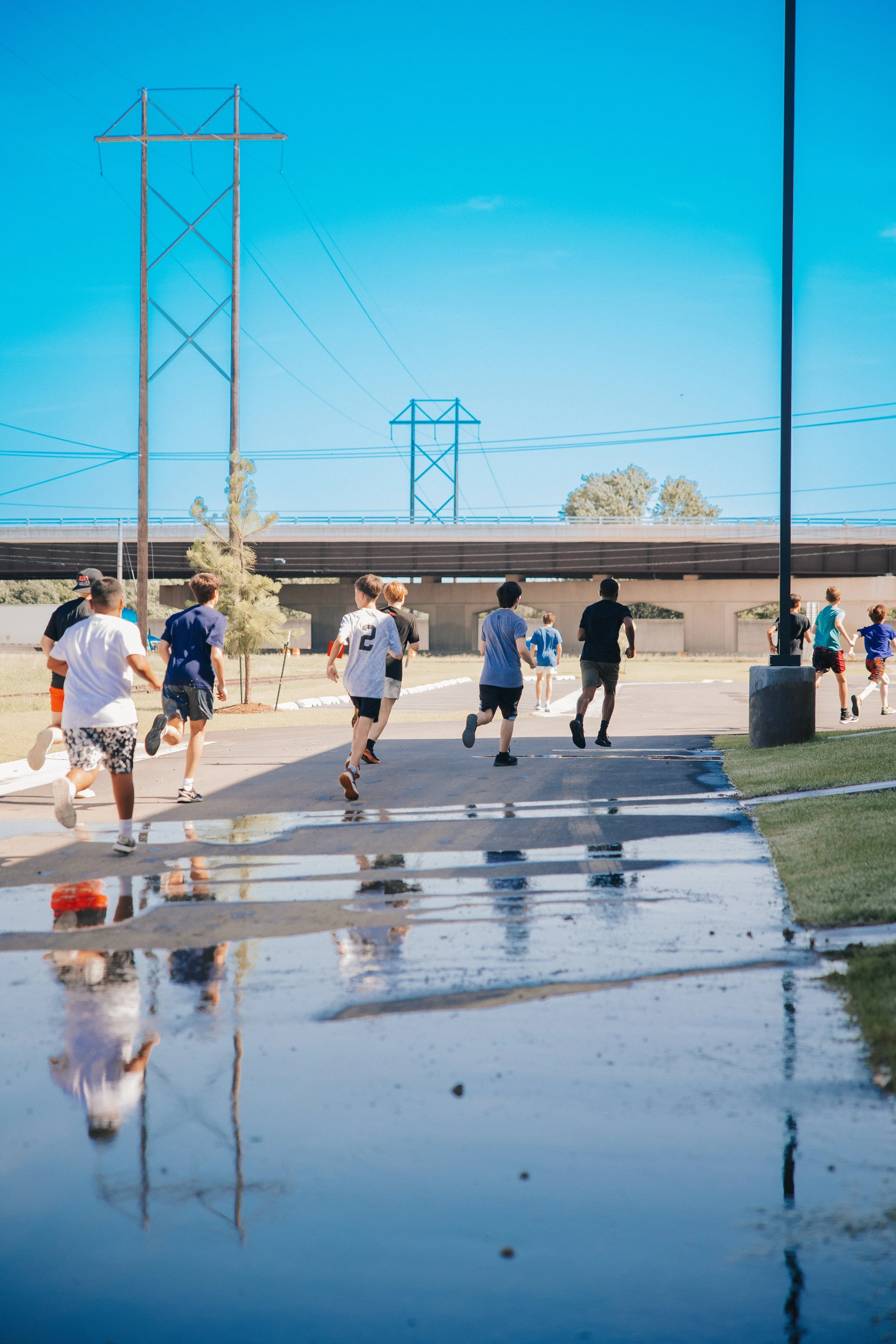Group of children running on a wet pavement with puddles, under a clear blue sky, near a bridge and power lines.