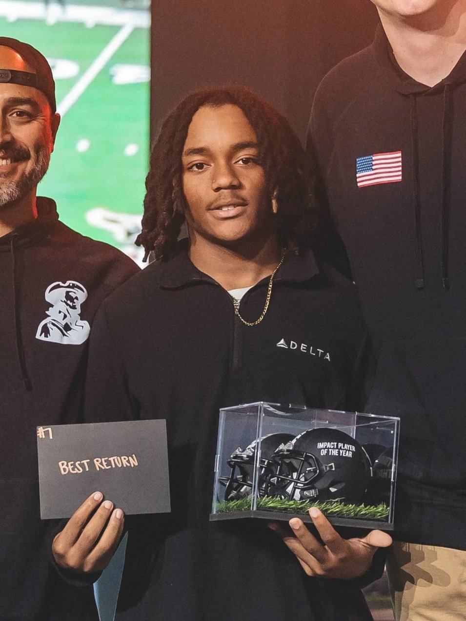 Young man with dreadlocks holding a clear display case with football helmets and a plaque reading 'Impact Player of the Year.' He is also holding a card that says 'Best Return.' He wears a black Delta jacket and has a gold chain around his neck.