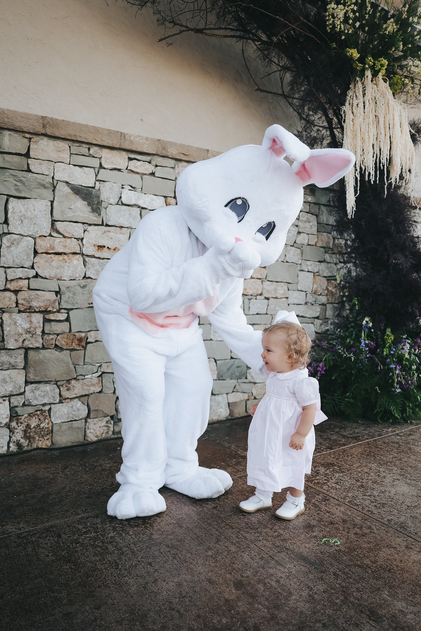 A person dressed in a white bunny costume with large ears and a cute face is kneeling and whispering to a young girl. The girl is wearing a white dress, white shoes, and a white bow in her curly blonde hair. They are outdoors near a stone wall with p
