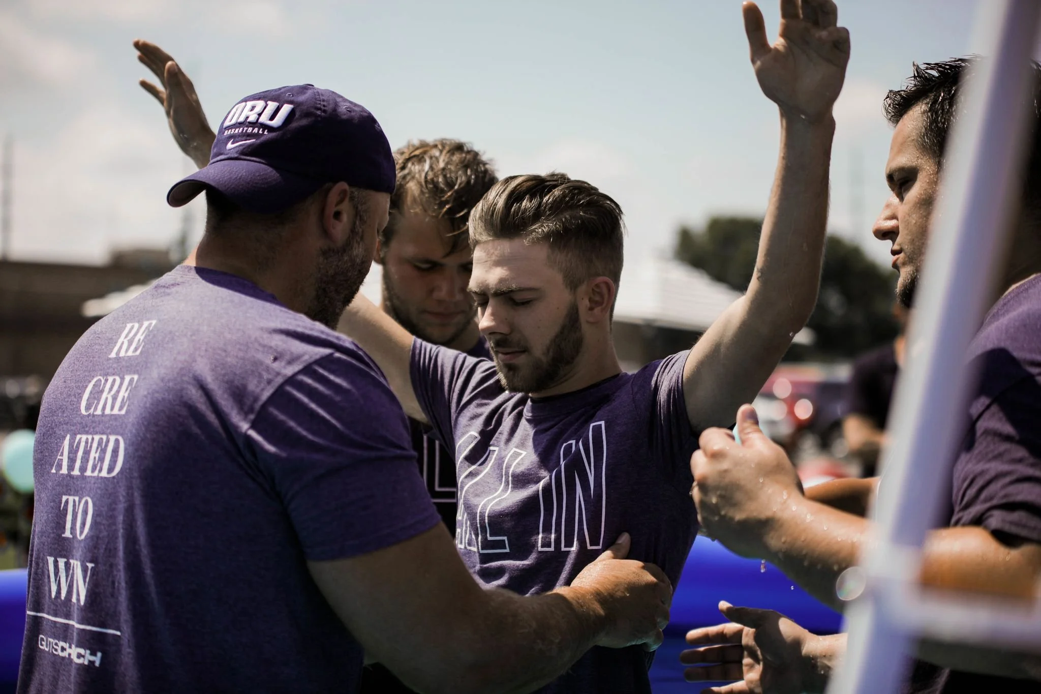 A group of men, some with water on their arms, are participating in a prayer or moment of reflection outdoors. One man in the center has his eyes closed and arms raised, while others surround and support him.
