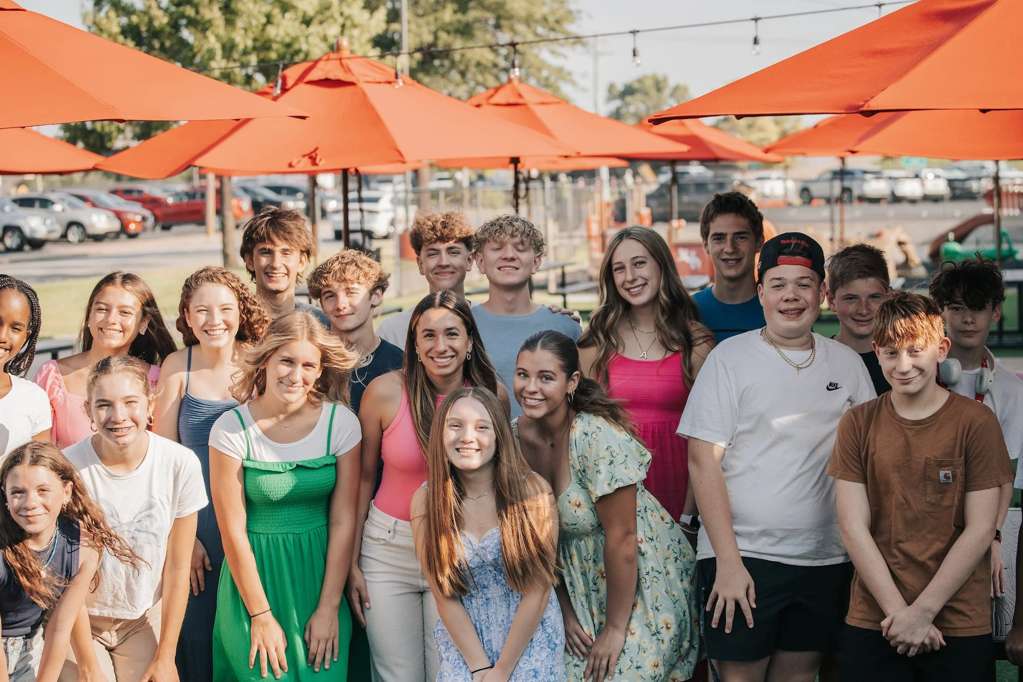 A group of teenagers smiling and posing for a photo outdoors under orange umbrellas at a park or recreational area, with cars and trees in the background.
