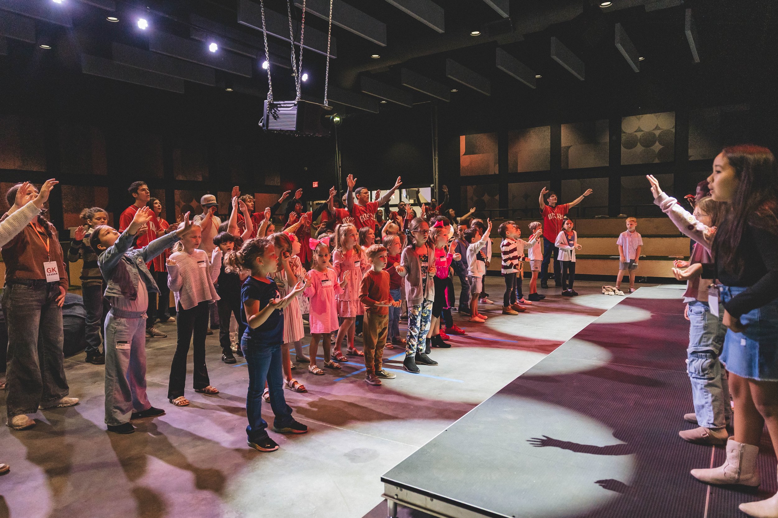 Children and adults participating in a group activity on a stage in an indoor auditorium, some with their arms raised, under stage lights.