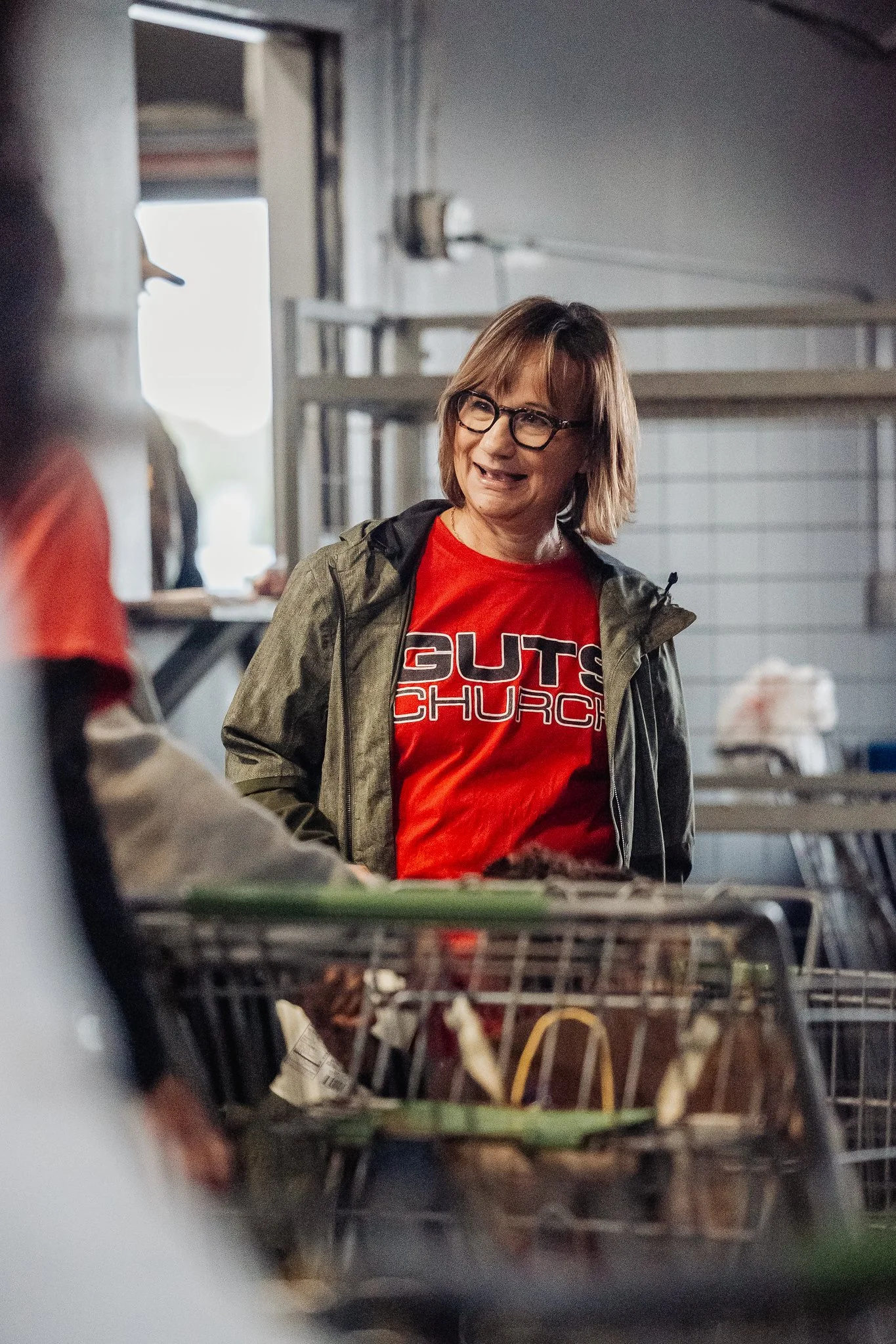 A woman wearing glasses and a red T-shirt with 'GUTS CHURCH' written on it, standing in a grocery store aisle near shopping carts.