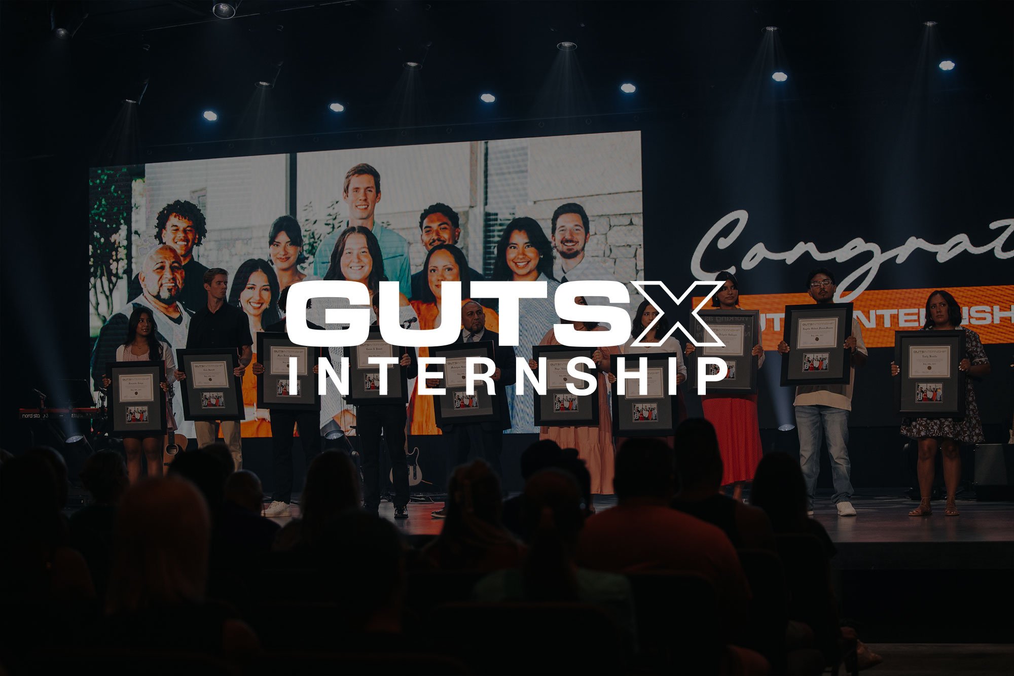 People standing on stage holding framed awards at a GUTS X Internship event, with a large screen behind them displaying a group photo and the words "Congratulations".