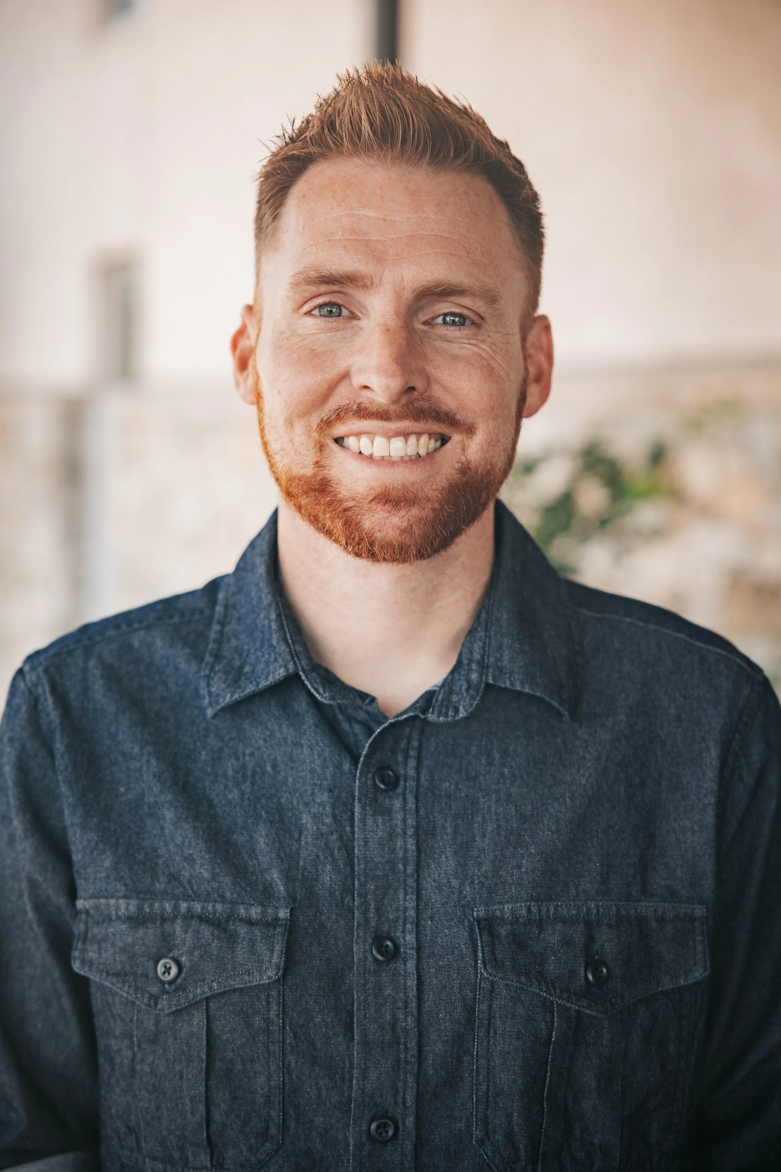 A man with red hair and a beard smiling, wearing a dark denim shirt, indoors with a blurred background.