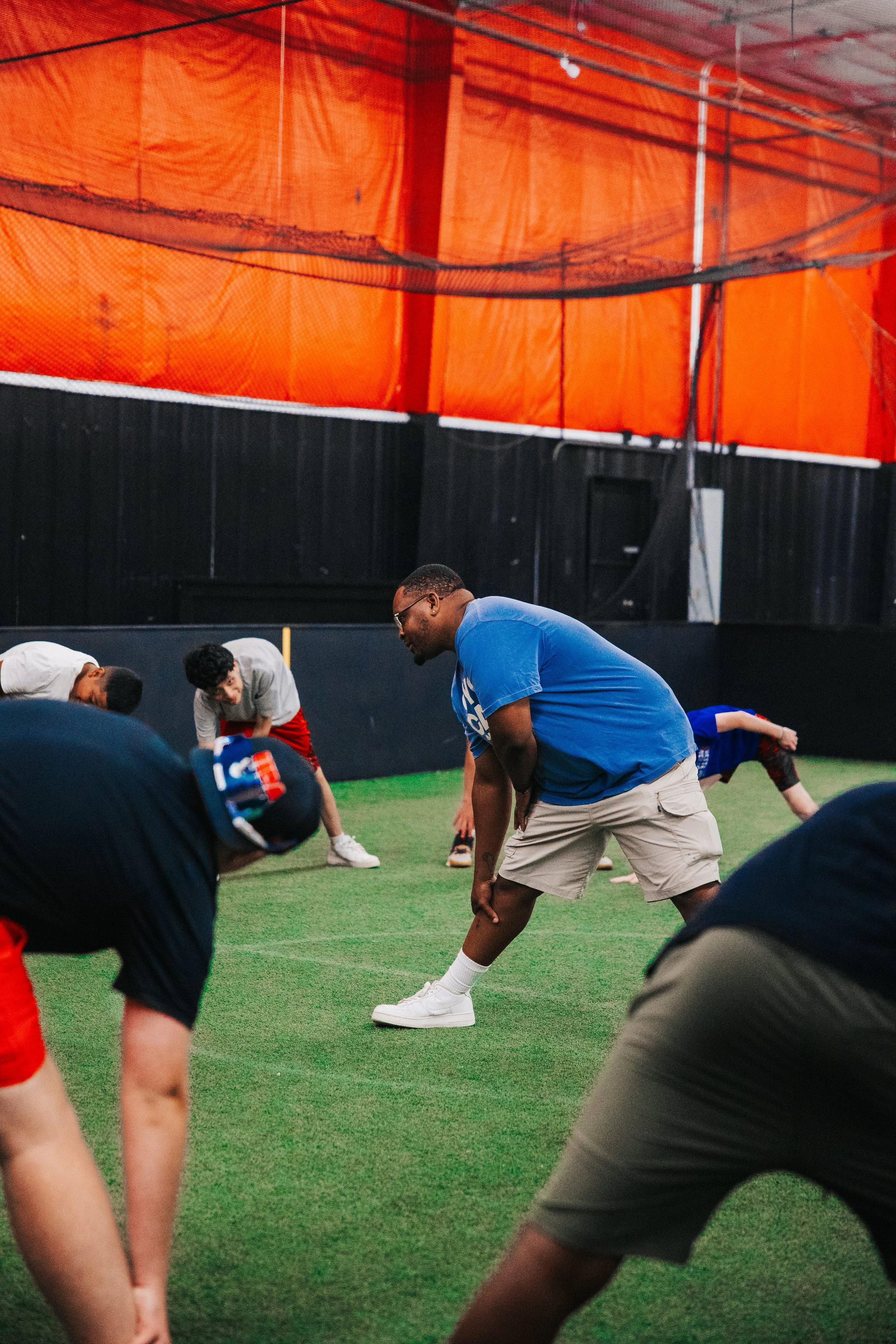 People participating in a stretching or exercise session indoors on a green turf field, with an orange and black background, and a coach or instructor leading the activity.