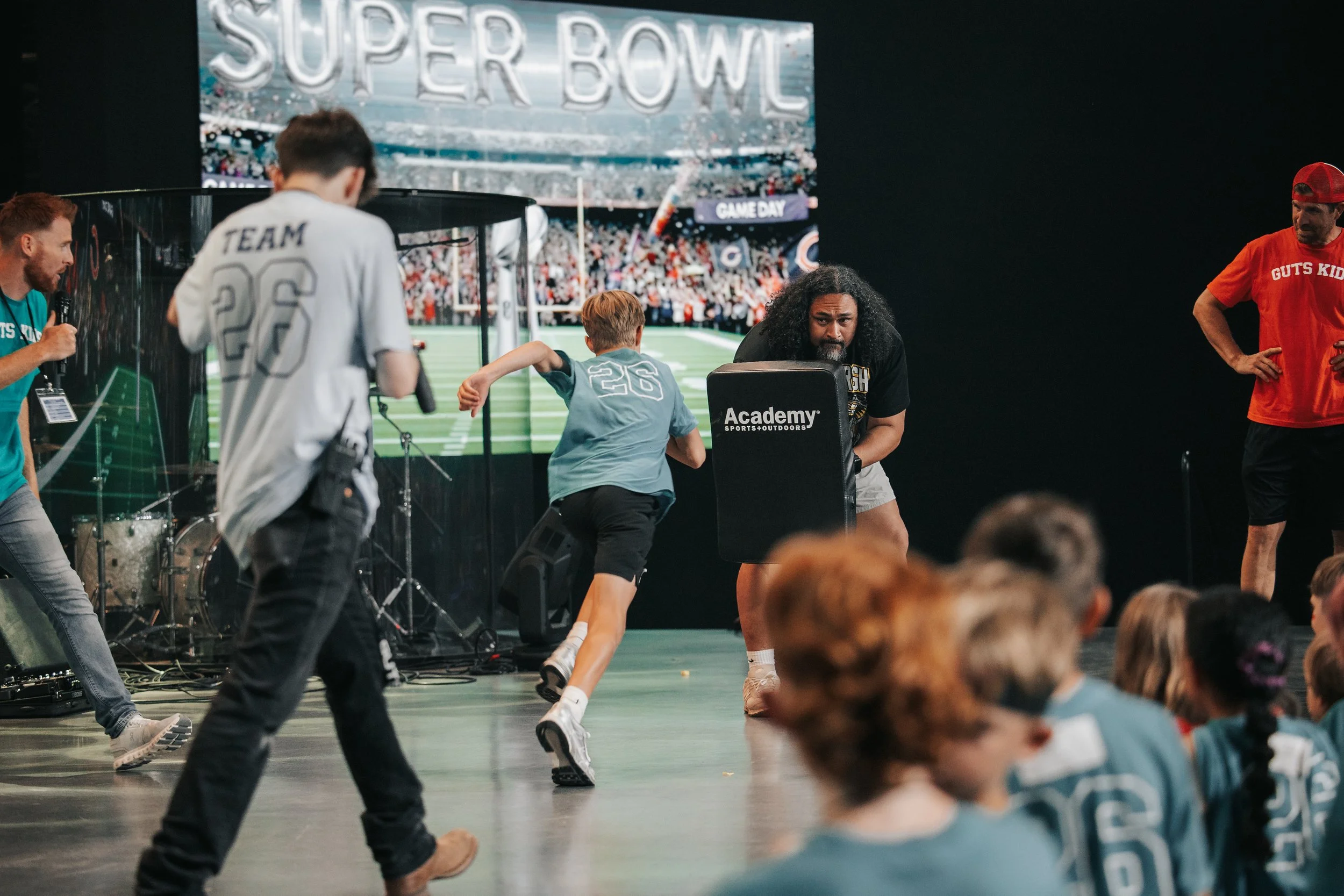 Kids playing a game at a Super Bowl event, with a large digital screen showing the Super Bowl logo in the background.