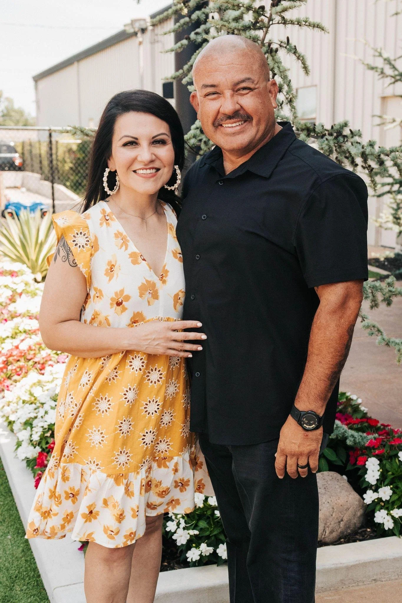 A smiling woman in a yellow floral dress and earrings standing next to a smiling man in a black shirt, outdoors in front of flowering plants and a tree.