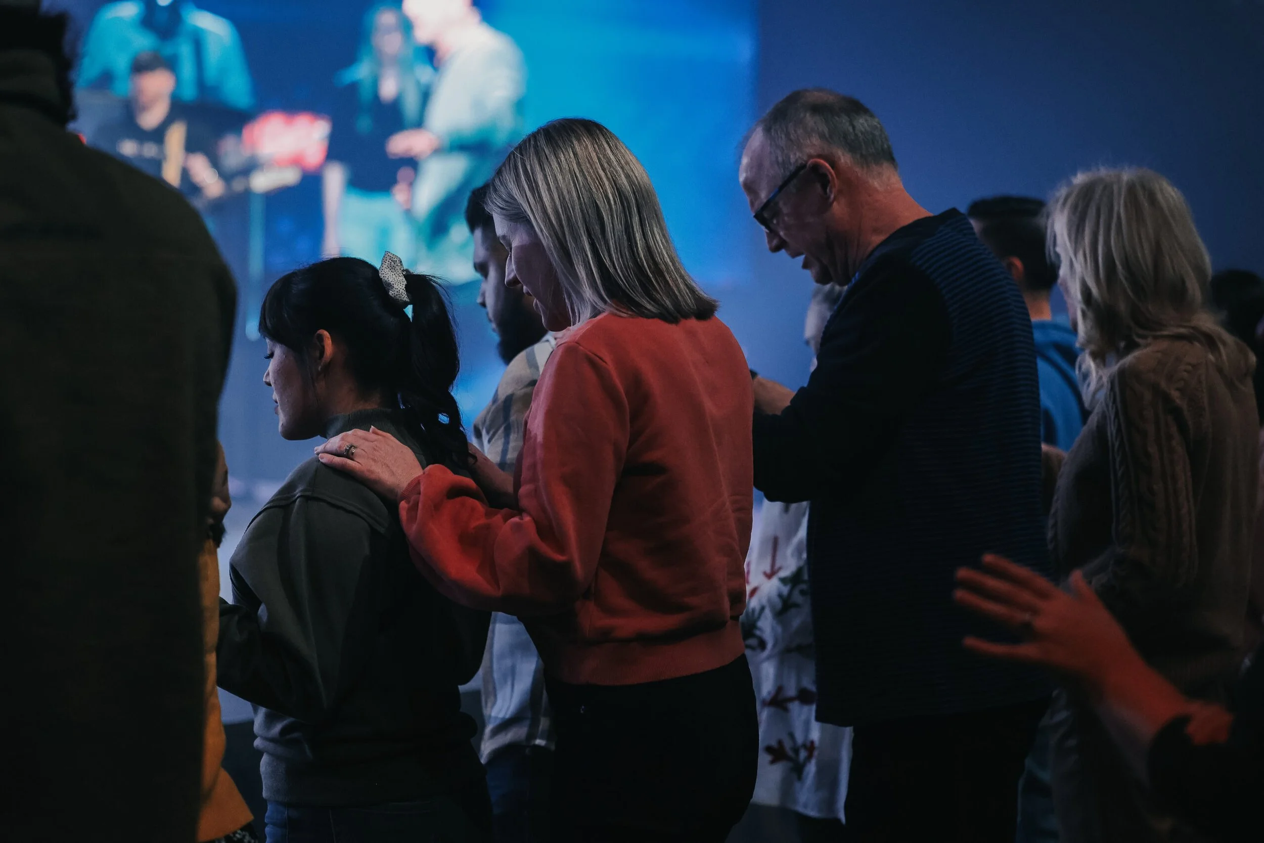 People praying with their heads bowed and hands on each other's shoulders, in a dark room with a blue-lit stage in the background.