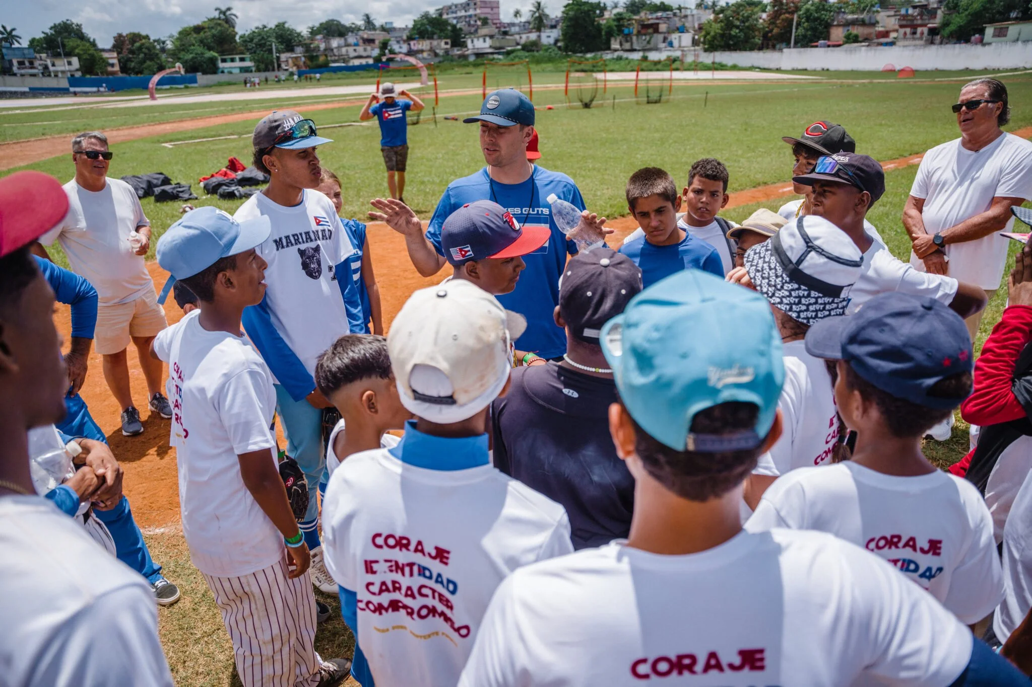 Group of young baseball players and coaches gathered on a baseball field during a team meeting or instructions, with a few adults present, and houses visible in the background, on a sunny day.