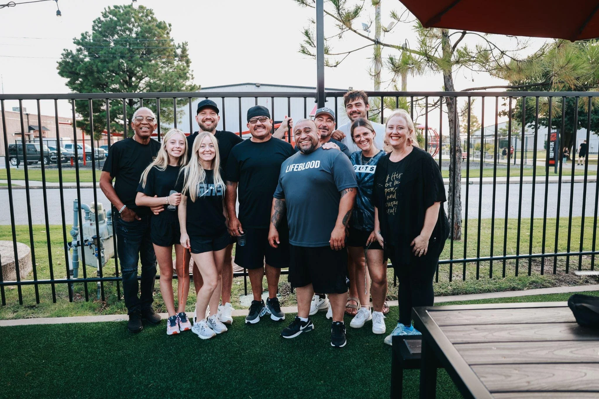 Group of men and women smiling in an outdoor setting, standing in front of a black fence, with trees, a parking lot, and a building in the background.