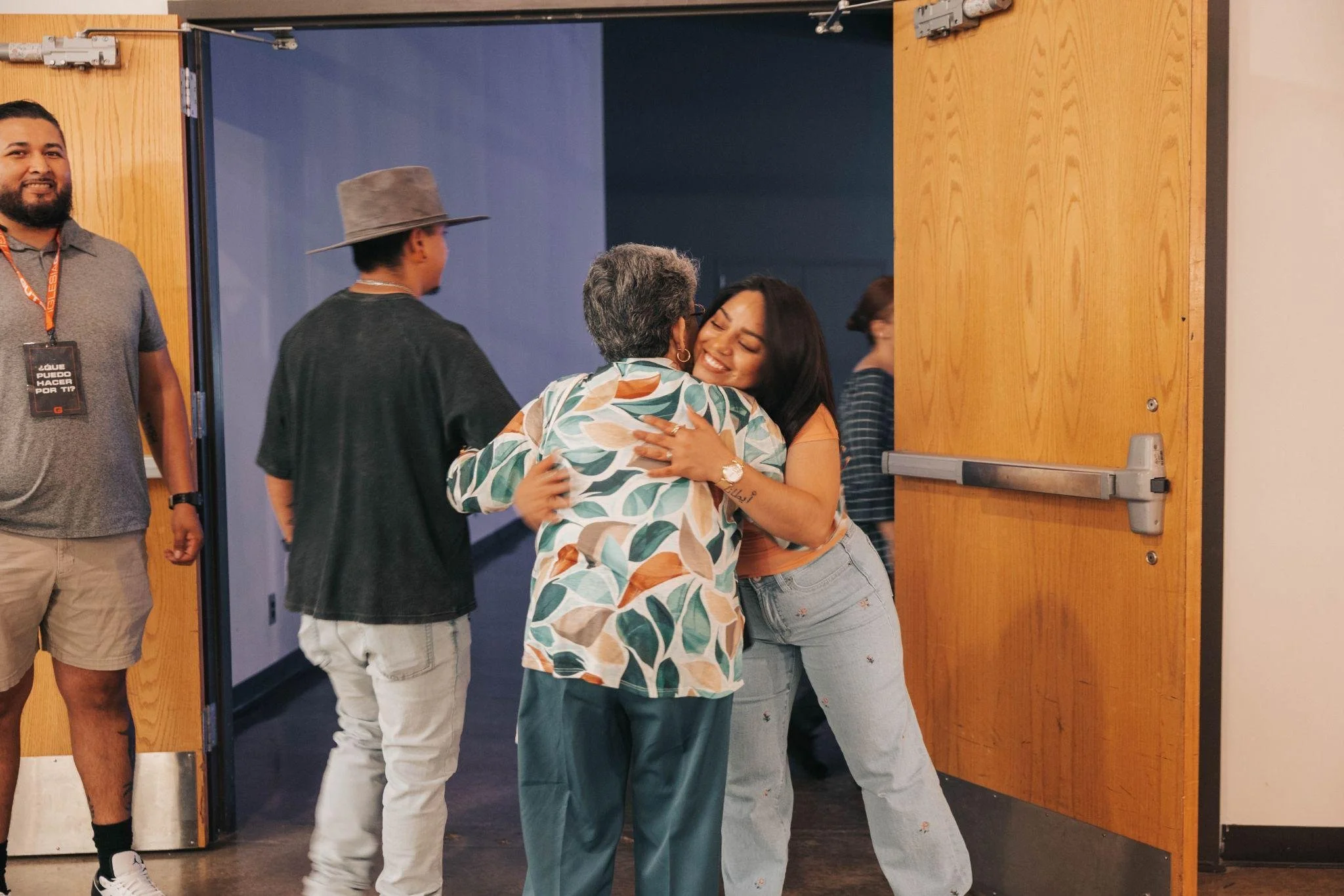 A woman with long black hair hugging an older woman in a patterned shirt at the entrance of a room, with other people in the background smiling and standing near the doorway.