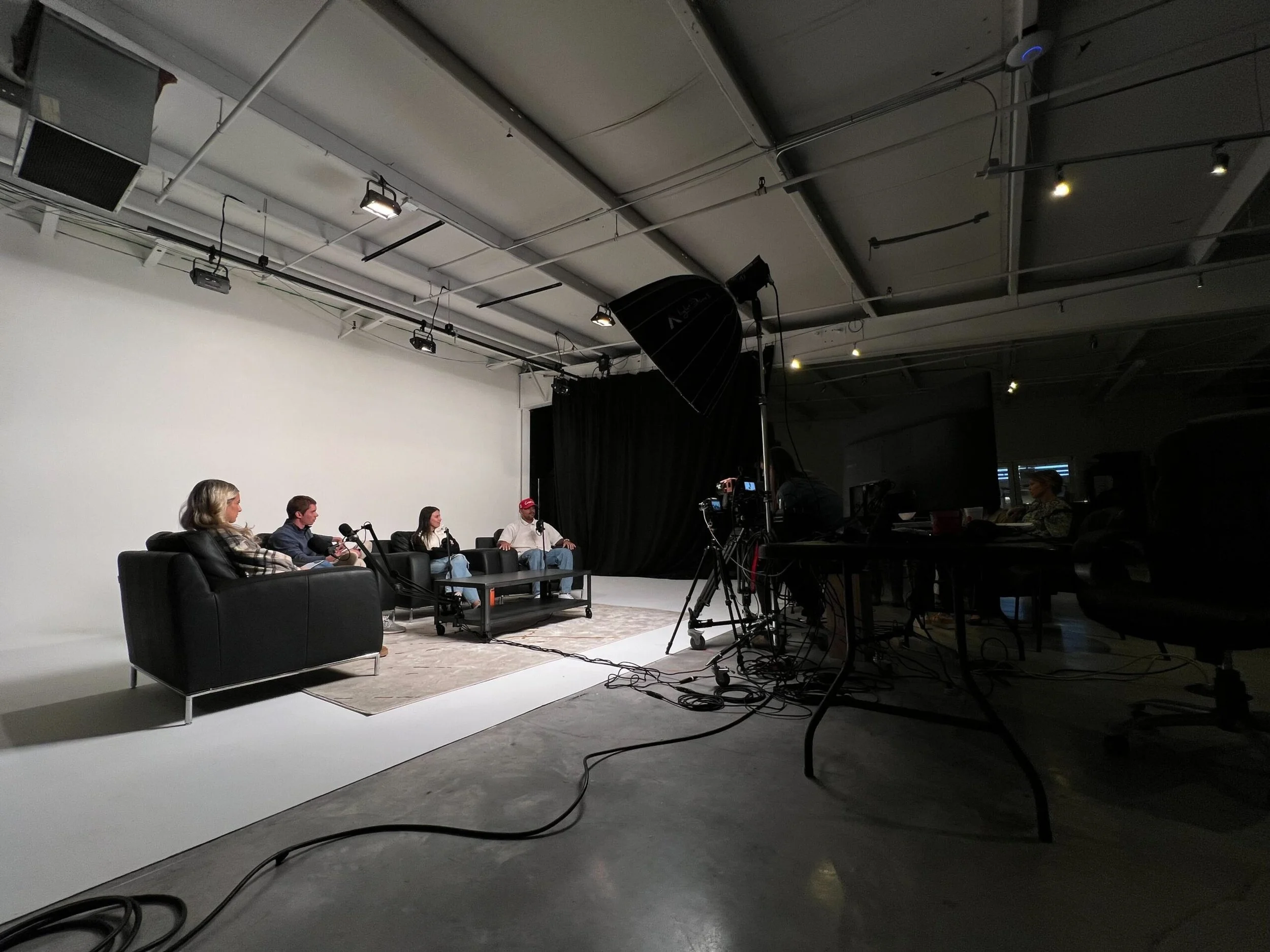 Four people sitting on black leather sofas in a studio, participating in a panel discussion with microphones. A camera and lighting equipment are set up in the foreground, with a plain white background and black curtains behind the panelists.