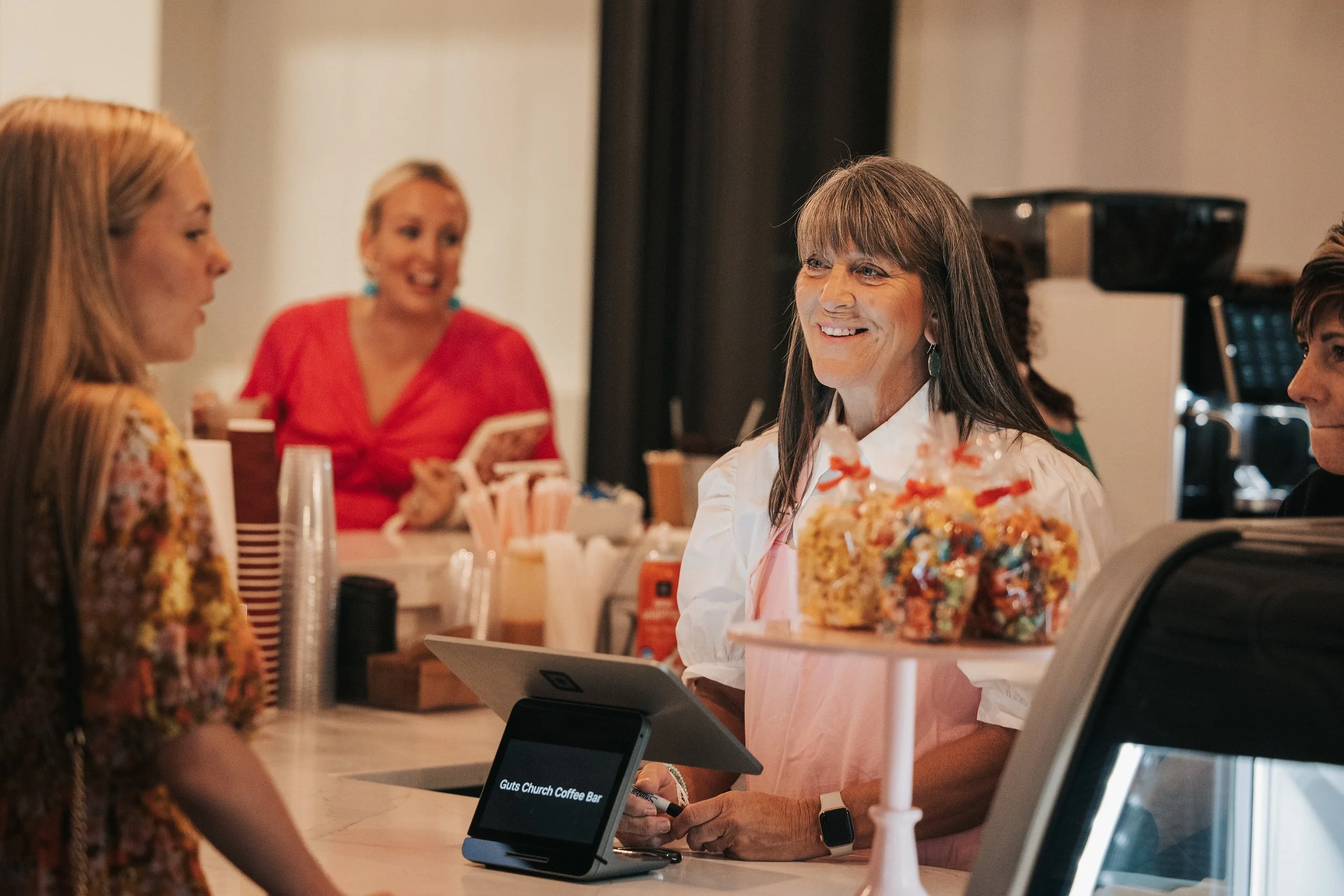 Women talking at a coffee bar, with one woman wearing a pink apron serving desserts, and others in casual clothing engaged in conversation.
