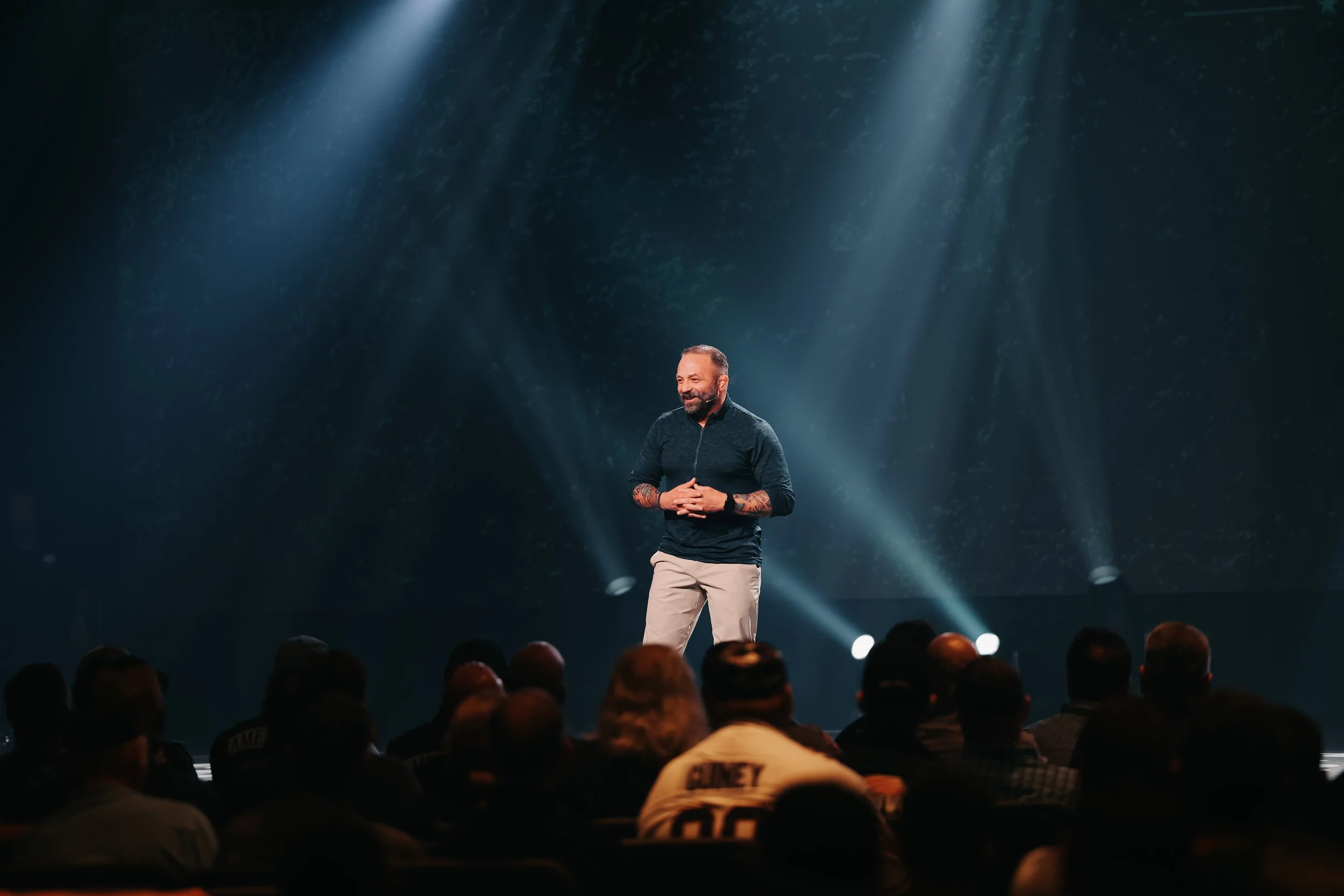 A man on stage speaking to an audience with a dark background and spotlight shines.